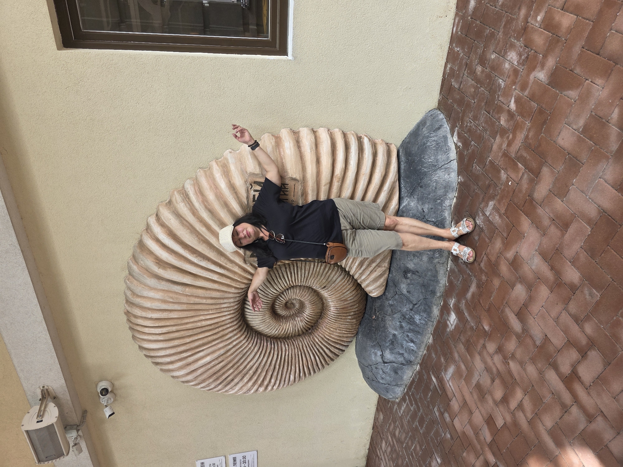 A woman stands in front of a large ammonite fossil replica in Estosadok, Russia. The fossil is a light brown spiral shape with detailed ridges, mounted on a dark grey rock base. The woman is wearing a beige baseball cap, a dark t-shirt, khaki shorts, and white sandals. She has a brown crossbody bag. Her right arm is extended towards the fossil, and her left arm is bent at the elbow. She appears to be posing for the photo. The background is a textured, light yellow wall with a dark brown framed window on the right side. Above the fossil, there are two security cameras and a white speaker mounted on a light grey beam. The ground in front of the fossil is paved with reddish-brown bricks laid in a herringbone pattern. Signs are visible on the wall to the left of the fossil.