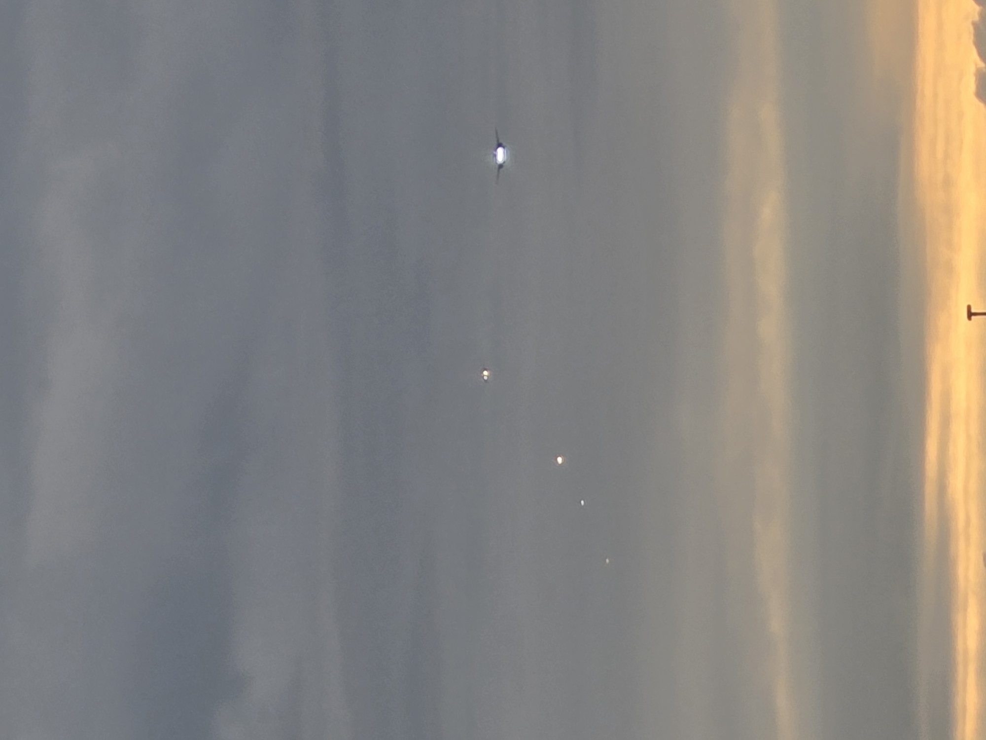 The image shows a cloudy sky above Kelsterbach, Germany, with a line of lights visible in the distance. The lights appear to be from aircraft or drones, moving in a diagonal formation from left to right. The sky is a mix of gray and yellow hues, suggesting it is either dawn or dusk. The bottom of the image shows a portion of what appears to be a street light.