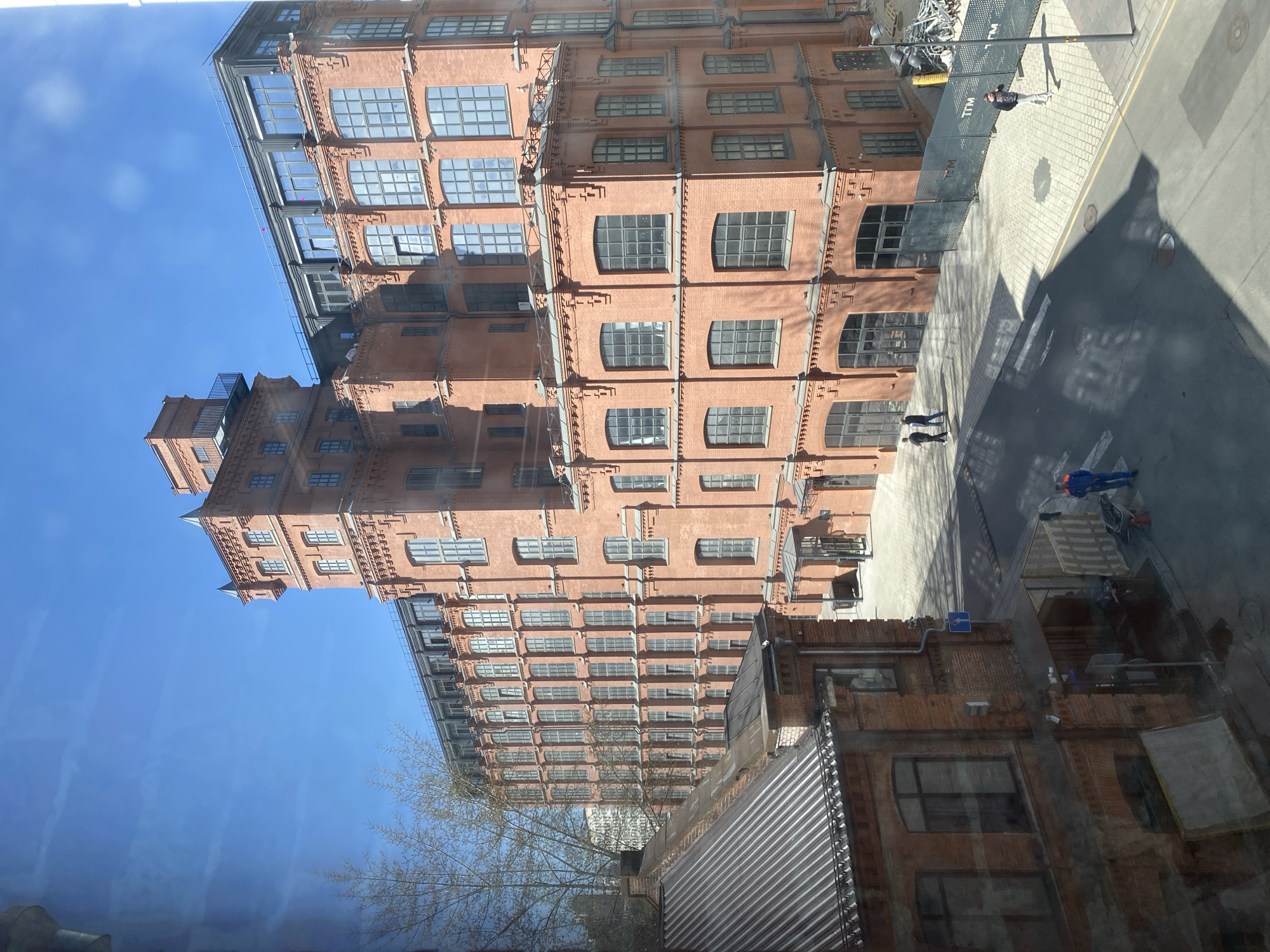 The image shows an exterior view of the Danilovskaya Manufactura, a historical industrial complex located in Moscow, Russia. The main subject is a large, multi-story brick building with rows of arched windows. The building appears to be undergoing renovations, with scaffolding visible on the upper floors. Several people are seen on the street-level area, including construction workers in blue overalls. The surrounding area is urban, with paved sidewalks and some trees visible. The sky is clear and blue. The image is taken from an elevated perspective, possibly from another building. There is a window reflection at the bottom, which may indicate that the picture was taken from inside of another building.