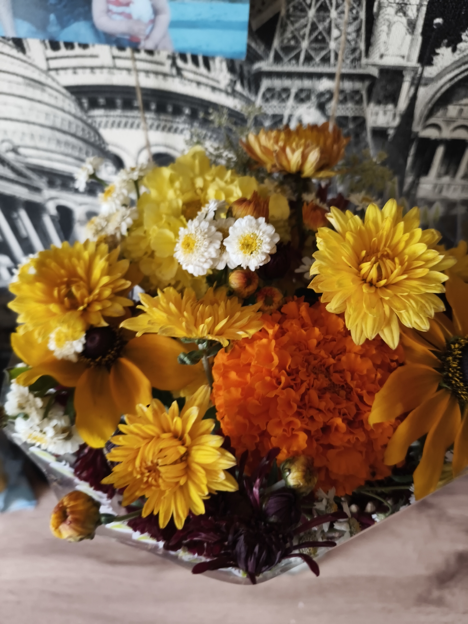 A close-up shot shows a bouquet of vibrant flowers, predominantly in shades of yellow, orange, and white, with hints of dark red foliage. The flowers are arranged in a way that suggests a festive or celebratory occasion. In the background, a tapestry or print depicts a stylized cityscape with iconic structures resembling the Eiffel Tower and other architectural elements, overlaid with a partially visible image of a child. The overall impression is one of warmth and cheerfulness. The image does not provide any information about the city of Zarudnya, Russia, or any specific location within it.