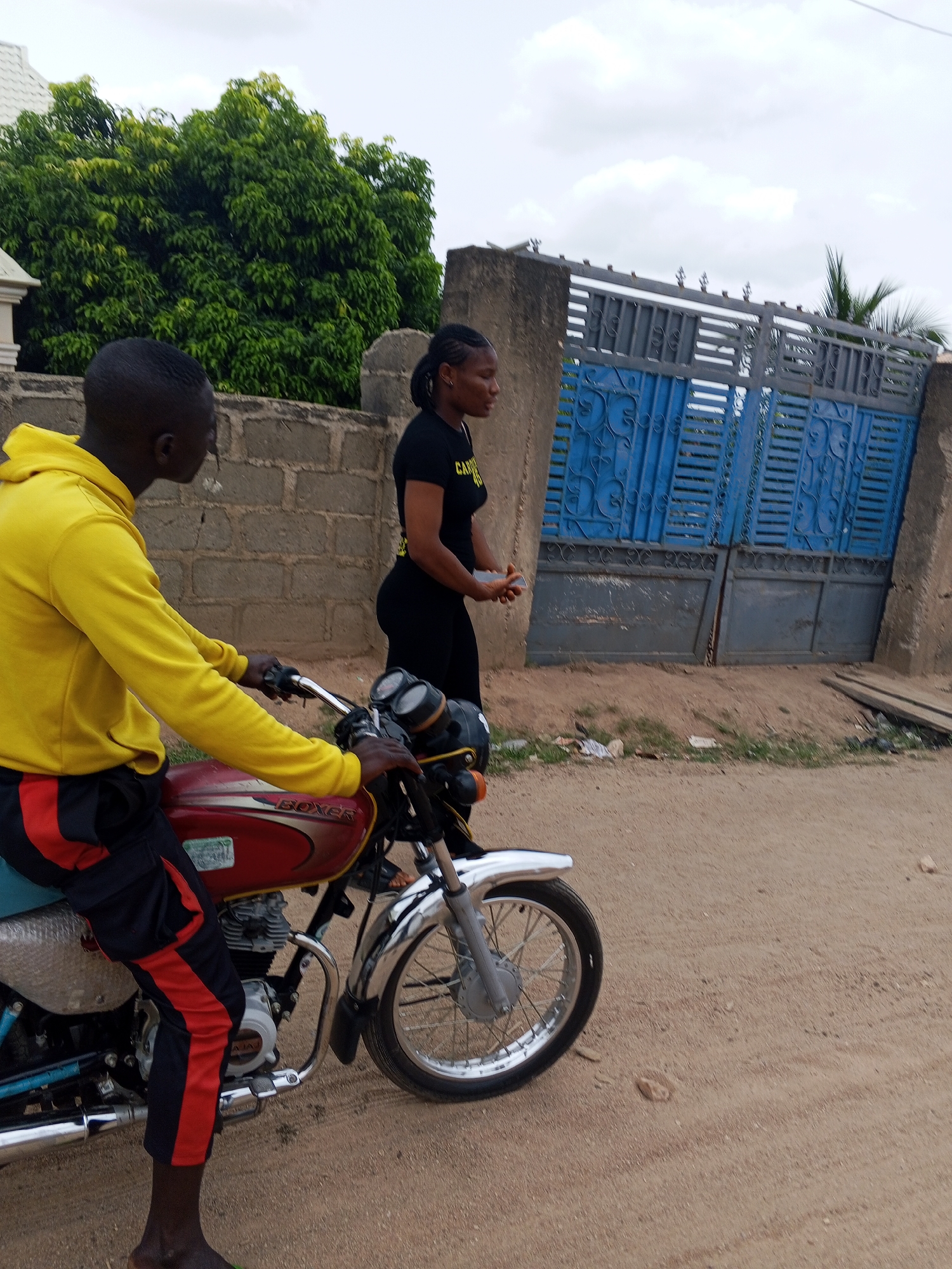 A person wearing a yellow hoodie and black pants with red stripes is sitting on a red Boxer motorcycle on a dirt road in Lokoja, Nigeria. Beside the motorcycle, a person in a black t-shirt and black pants is standing. Behind them are a brick wall, a large tree, and a blue and gray metal gate. The sky is overcast.