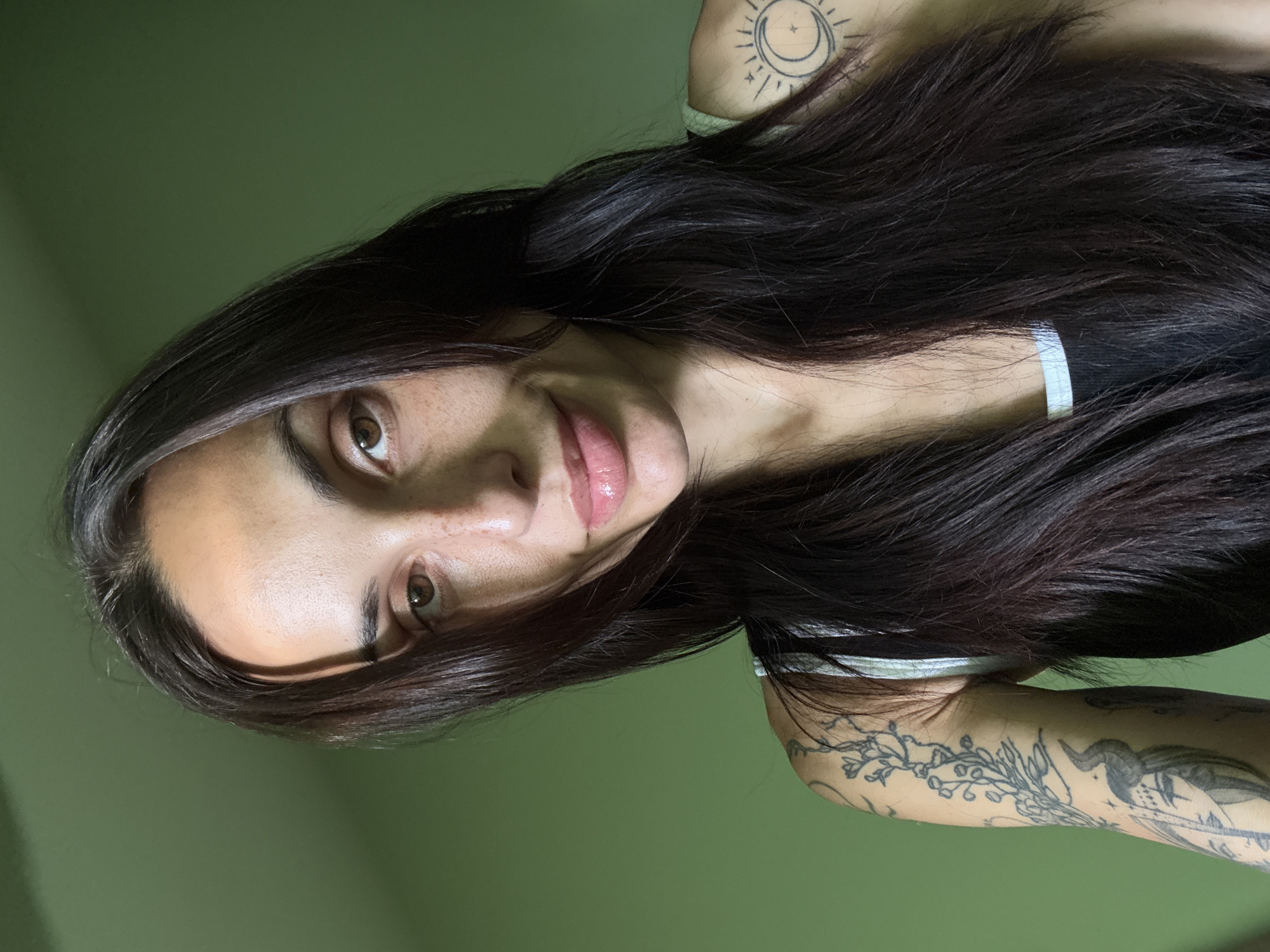 A woman with long, dark hair is looking at the camera. She has brown eyes and fair skin. She has tattoos on both arms, one with a sun and moon design, and another with vines and a horned figure. She is wearing a black and white tank top. The background is a solid green color. There is no indication of the location being Pleven, Bulgaria.