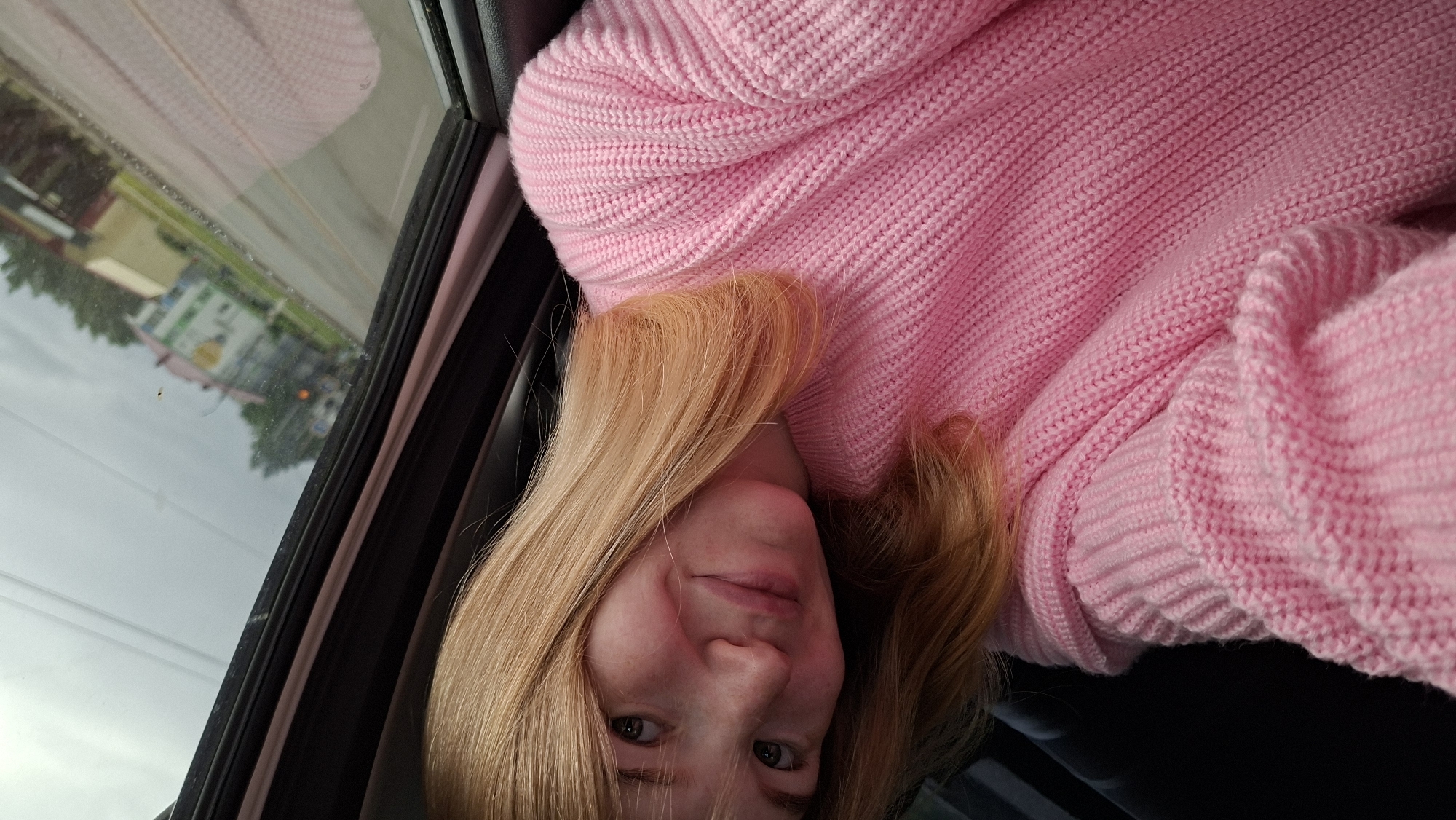 A young woman with blonde hair and fair skin, wearing a pink knitted sweater, is looking at the camera from inside a vehicle. Her head is tilted, and she has a slight smile. The background visible through the window shows a street with buildings and trees, suggesting an urban or suburban setting in Chaikovsky, Russia, under an overcast sky. The interior of the vehicle is dark, with the window frame and a portion of a dark seat visible.
