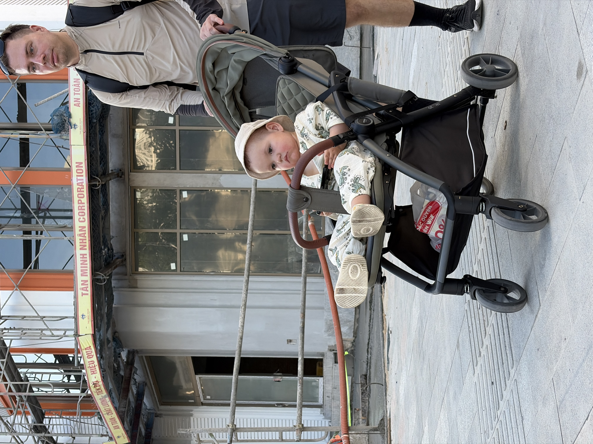 A man and a baby in a stroller are captured on a paved sidewalk in an urban setting. The man, on the left, looks directly at the camera with a neutral expression, wearing a light-colored long-sleeved top, dark shorts, and sunglasses on his head. He has a dark shoulder bag. The baby, seated in a grey stroller with a brown handlebar, also looks towards the camera with a curious expression. The baby wears a light patterned romper and a matching bucket hat, with visible white-soled shoes.
In the background, a building appears to be under construction or renovation, featuring scaffolding and large windows. A prominent yellow and red banner stretches horizontally across the mid-ground. The ground is light grey paved tiles, some with tactile strips. Bright daylight illuminates the scene.
Visible text on the yellow banner includes "AN TOÀN - CHẤT LƯƠNG TIẾN DŨ - HIỆU QUẢ" (Safety - Quality - Progress - Efficiency) and "TÂN MINH NHÂN CORPORATION". Fragmented text like "ĐẶC QUYÊN" and "EN WIN" is seen on a plastic bag stored under the stroller, where "AN TOÀN" is also partially visible on a label. The location context is Đà Nẵng, Vietnam.