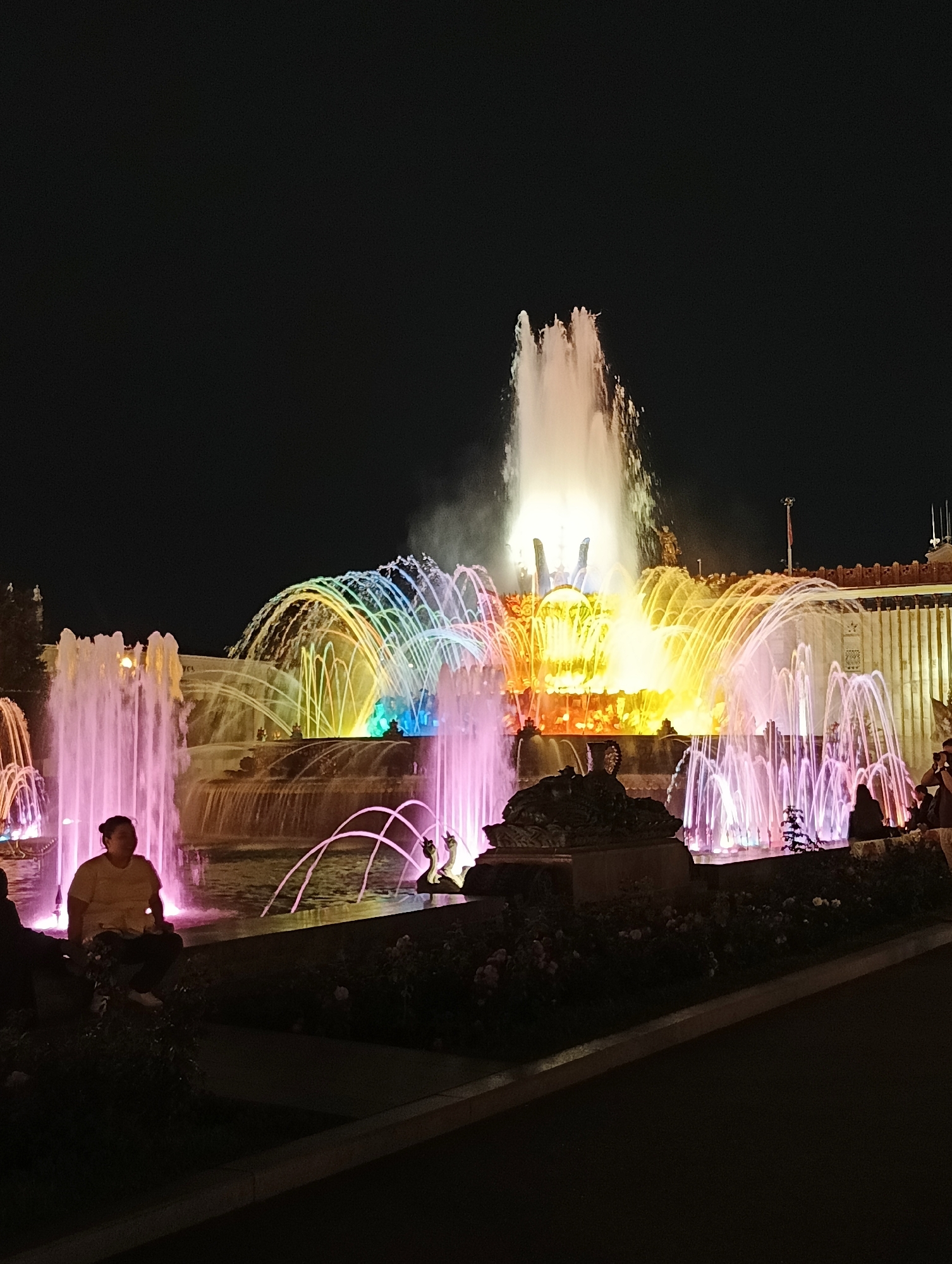 The image shows the Friendship of Peoples fountain at night, illuminated with colorful lights. The large, central fountain features a towering water spray, while surrounding fountains emit arcs of water lit in pink, purple, yellow, and rainbow hues. Several people are visible sitting on the edge of the fountain or in the foreground, silhouetted against the light. The background shows the illuminated facade of a building, likely part of the All-Russia Exhibition Centre (VDNKh). The scene captures a vibrant and lively atmosphere, with the illuminated fountains serving as the main attraction.