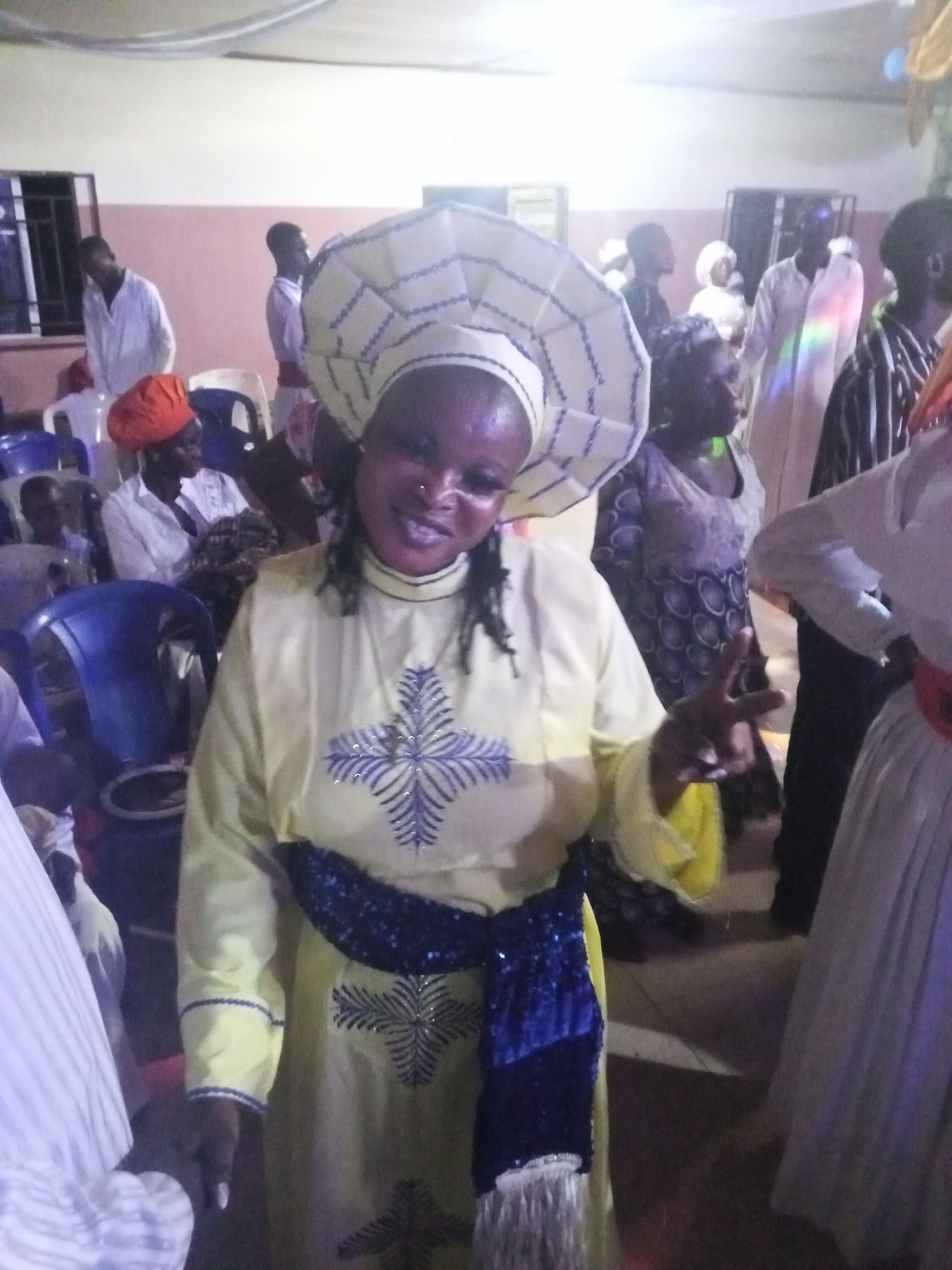 The photograph shows a woman in a light yellow dress with blue embroidered designs and a matching blue sash. She is wearing a large, white hat with blue trim. She is smiling and holding up a peace sign with her left hand. The scene appears to be inside a building with a number of people in the background, dressed in similar attire. The setting is likely a religious gathering or ceremony, possibly in the city of Lokoja, Nigeria. There are chairs and other people in the background, some wearing white garments, and a man in an orange hat can be seen in the distance.