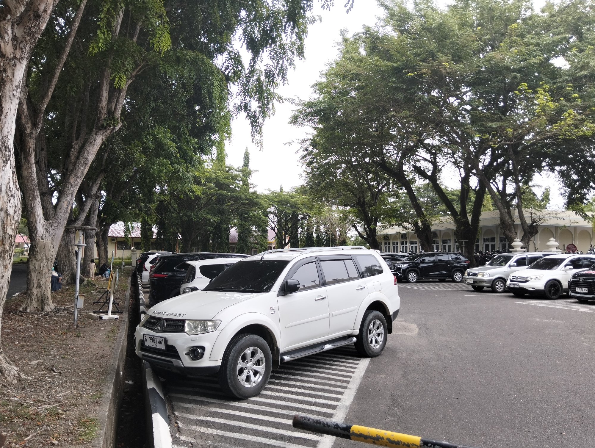 A white Mitsubishi Pajero Sport SUV is parked on a paved lot, angled towards the right of the frame. The SUV is facing slightly away from the viewer, with its front right tire visible. The vehicle has tinted windows, silver roof rails, and chrome accents. A black and yellow striped barrier is in the foreground, partially obscuring the lower right portion of the image. The parking lot is lined with several other vehicles, including black SUVs and a silver SUV. In the background, a building with white columns and a red tiled roof is visible, partially obscured by lush green trees. Several large trees with rough bark line the left side of the image, casting shadows on the ground. The ground is covered in dirt and fallen leaves. Two individuals are visible in the middle ground, to the left of the parked SUVs, near a pole. One appears to be standing and wearing dark clothing, while the other is crouched down. The overall scene suggests a parking area in a semi-urban or institutional setting in Tambontunong, Indonesia, possibly a government building or university campus given the architecture and manicured trees. The sky is overcast.