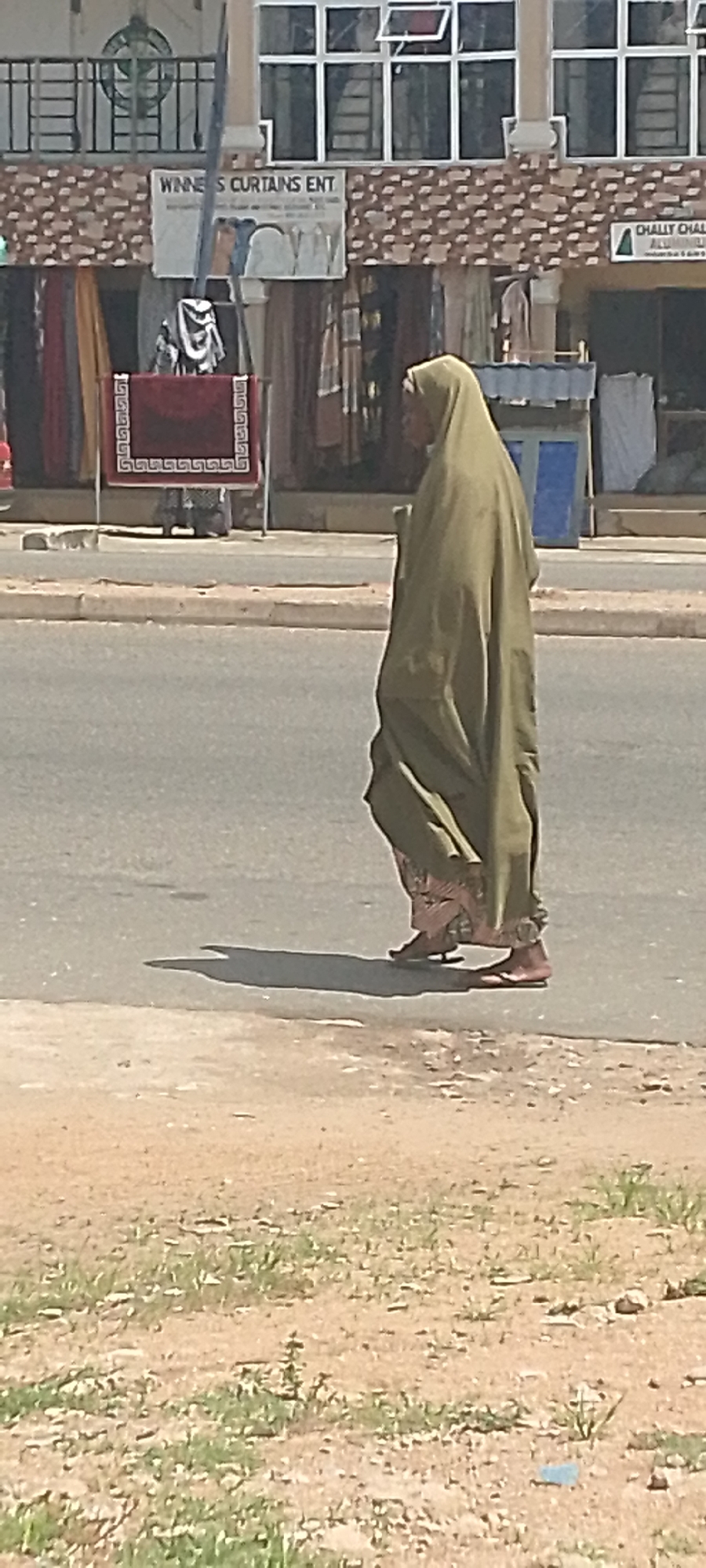 A person, wearing a light green headscarf and a matching abaya, walks across a paved road. The person's face is not clearly visible, as they are seen in profile from the right side of the frame, looking to the left. They are wearing sandals. A dark shadow is cast on the road by the person, suggesting it is daytime.
Behind the person, there is a street lined with shops. A banner with the text "WINNERS CURTAINS ENT" is visible on one of the shops. Another banner further to the right has the text "CHALLY CHAL". Colorful textiles, possibly curtains or fabrics, are displayed in front of the shops. The setting appears to be a market or commercial area. The ground in the foreground is unpaved and covered with sparse vegetation. The overall impression is of a sunny day in a bustling urban environment.
