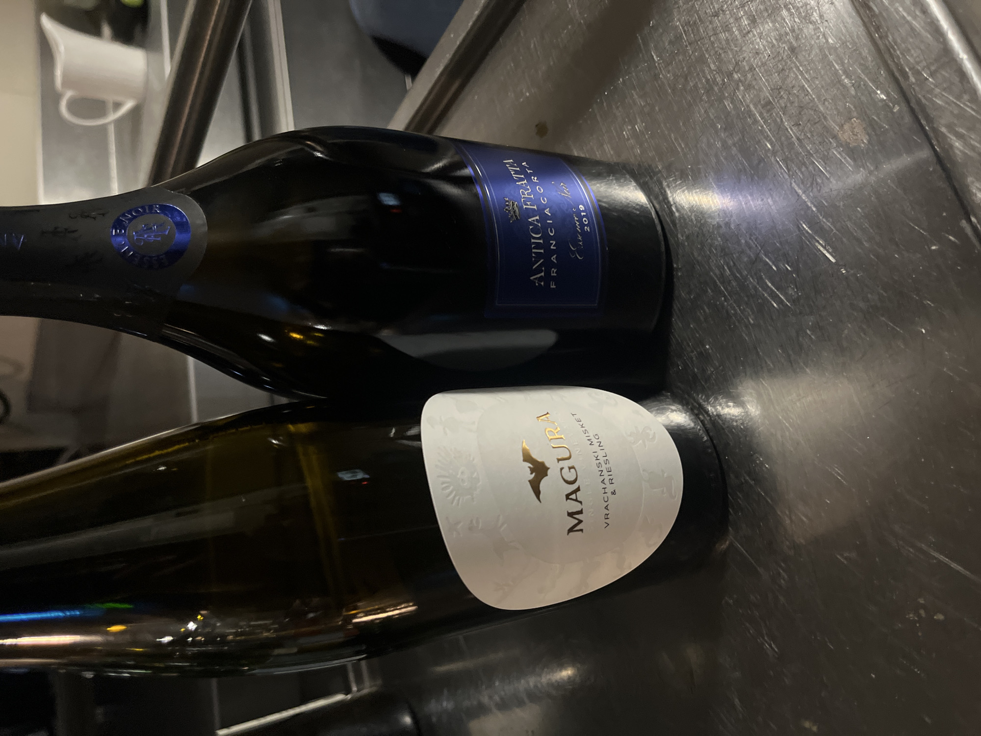 The image shows two wine bottles lying on a stainless steel surface. The top bottle has a blue label that reads "ANTICA FRATTA FRANCIACORTA" and the year "2019". The bottom bottle has a white label with the word "MAGURA" in gold lettering, followed by "VRACHANSKI MISKET & RIESLING". The photo was likely taken in a kitchen or bar area, as evidenced by the stainless steel surface. The location appears to be in Bulgaria, specifically the city of Sofia, due to the presence of a bottle with Vrachanski Misket, a type of wine typically produced in Bulgaria.