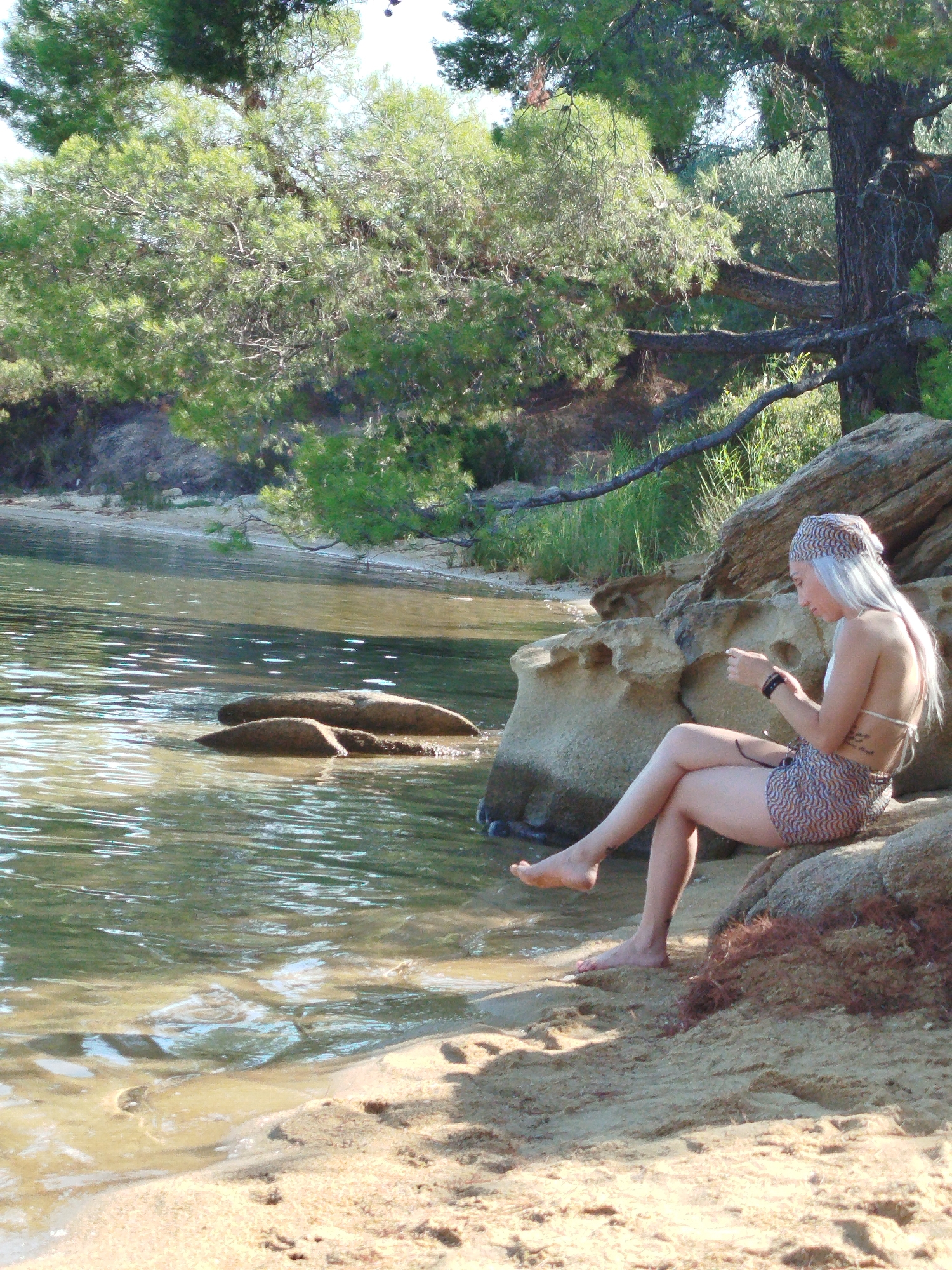 A young Caucasian woman with long, pale blonde hair is sitting on rocks by the sea. She is wearing a patterned bandeau bikini top and matching patterned shorts, along with a matching headscarf. She is looking down at her hands, which are holding a mobile phone. The woman is barefoot and has a tattoo on her left thigh. The sea is calm and shallow, with the sandy bottom visible in places. There are rocks and vegetation in the background, with trees overhanging the water. The scene is set in a natural, outdoor environment, likely a beach or cove. This appears to be a serene moment of relaxation. The location is the Sithonia Municipal Unit, Greece.