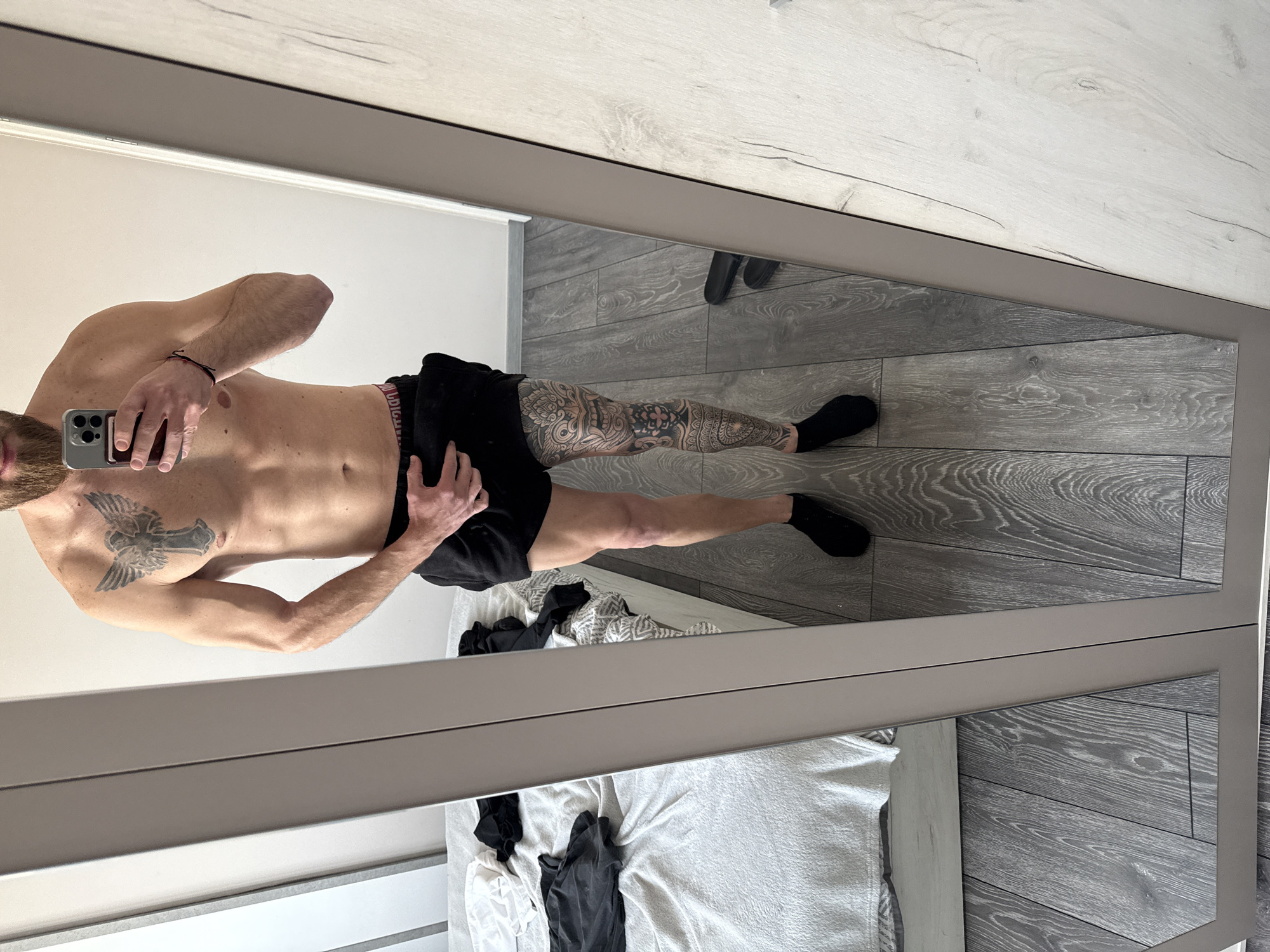 A man is reflected in a full-length mirror. He is facing the mirror and taking a photo of himself with his mobile phone. He is shirtless, wearing black shorts, and black socks. He has a tattoo on his left chest and his right leg is heavily tattooed. The room contains a bed with white sheets and clothing scattered on it, as well as grey wooden floors. It is not possible to determine the location in Sofia, Bulgaria from the image.