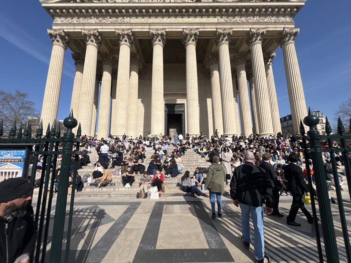 The image depicts the front facade of the Panthéon in Paris, France, under a clear blue sky on a sunny day. The neoclassical monument features a prominent colonnaded portico with Corinthian columns and a pediment bearing the inscription "AUX GRANDS HOMMES LA PATRIE RECONNAISSANTE." A substantial crowd of individuals, predominantly young adults, are gathered on the wide stone steps leading up to the main entrance. Most are seated casually across the steps, in various postures, some appearing to converse, others looking forward or at personal devices. Several individuals are actively walking up or down the steps.
In the foreground, a dark green metal fence with decorative, spiked finials partially frames the scene. The ground immediately in front of the fence consists of a patterned surface with alternating dark and light gray striped and square tiles. To the far left foreground, a man with a dark beard and a black head covering, possibly a turban, is partially visible, holding a light-colored cup in his hand. Near the center-right foreground, a man wearing a black jacket and blue jeans walks up the steps, away from the camera. Alongside him, another man in a dark suit is also walking up, carrying a small yellow object in his right hand. Bare-branched trees are visible on either side of the Panthéon, contrasting against the bright sky, and distinct shadows are cast by the monument and people, indicating direct sunlight.