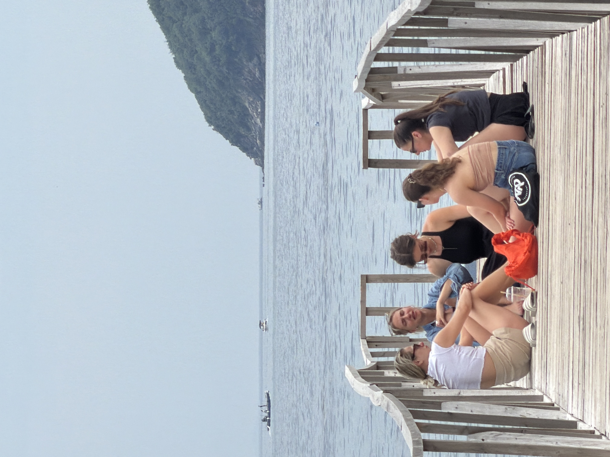 The image shows four women sitting on a wooden dock in Budva, Montenegro. The dock overlooks a body of water with a mountain range in the background. One woman is wearing a black shirt and black shorts. Another woman is wearing a tan top and denim shorts with a black logo patch. The third woman is wearing a black tank top. The fourth woman is wearing a blue denim shirt. The fifth woman is wearing a white t-shirt and tan shorts. An orange tote bag is on the dock.