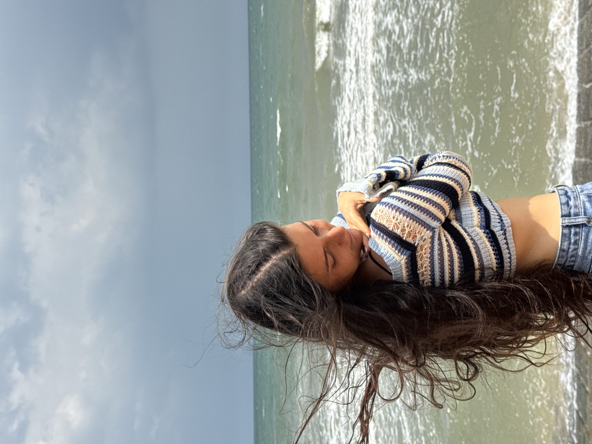 A young woman with long, dark, wavy hair stands on a paved surface by the ocean. She is wearing a striped, cropped sweater and denim shorts. Her head is bowed and her eyes are closed as she rests her head on her arms, her hair blowing in the wind. The ocean is choppy with waves and the sky is overcast. The location is not identifiable as Phu Thuy Ward, Vietnam.