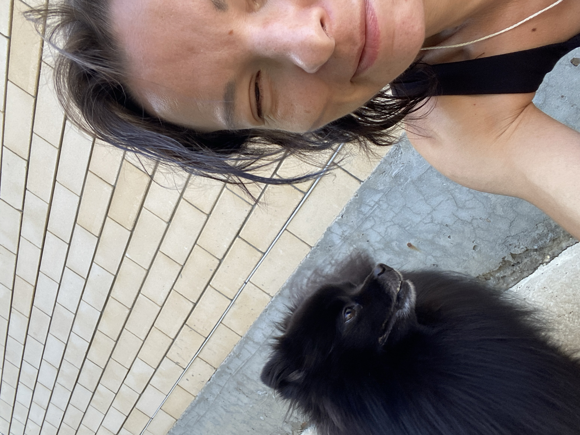 A close-up selfie shows a woman with dark hair and a black tank top, smiling with one eye closed, next to a small black dog with fluffy fur. The background consists of a textured, light-colored tiled wall and a rough concrete surface. The image is taken outdoors, possibly in Yuzhny, Russia, as suggested by the provided context. The woman's expression appears relaxed and happy as she poses for the photo with her pet. The dog is looking to the side, with its head tilted slightly.