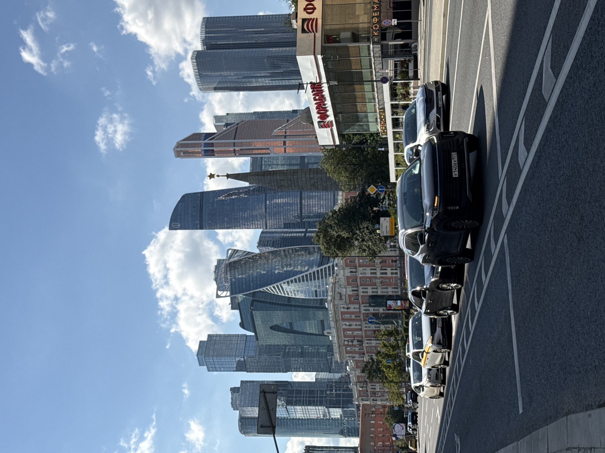 The image captures a street scene in Moscow, Russia, with a prominent view of the Moscow International Business Center, also known as Moscow City. The foreground features a multi-lane road with several cars in traffic, including a black SUV with license plate Y140BX 977, a silver sedan, and several other vehicles partially obscured. The road has clear lane markings and a pedestrian sidewalk on the left.
In the middle ground, a low-rise building with a white facade and red accents stands, housing businesses with visible signage. One sign displays "ФОРА-БАНК" (Fora-Bank) in red lettering. Adjacent to it, another sign reads "КОФЕМАНИЯ" (Kofemania), a Russian coffeehouse chain. Trees line the street in front of this building.
The background is dominated by the distinctive skyscrapers of Moscow City, showcasing a variety of modern architectural designs. Notable among them is a tall, dark building with a star at its peak, and other glass-facade towers with unique geometric shapes. The sky above is blue with scattered white clouds, indicating a clear day. The overall impression is a bustling urban environment with a mix of contemporary architecture and everyday city life.
