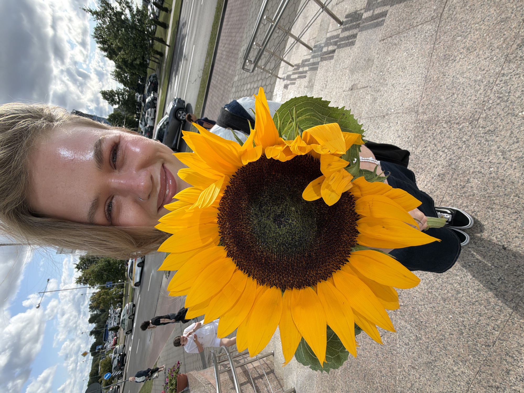 A young woman with blonde hair and a nose ring is holding a large sunflower in front of her. She is smiling and looking at the camera, her face illuminated by the sun, which also creates a shiny sheen on her forehead and cheeks. She is wearing a dark top and dark pants, with black and white Vans sneakers. The sunflower has vibrant yellow petals and a dark brown center, and its stem is visible. The woman is standing on granite stairs in an outdoor setting. In the background, there is a street with traffic, including cars and a bus. Trees line the street, and a cloudy sky with blue patches is overhead. A few other people are visible in the background, walking on the sidewalk and near the street. A metallic railing runs along the stairs. The location is identified as Бараўлянскі сельскі Савет, Belarus.
