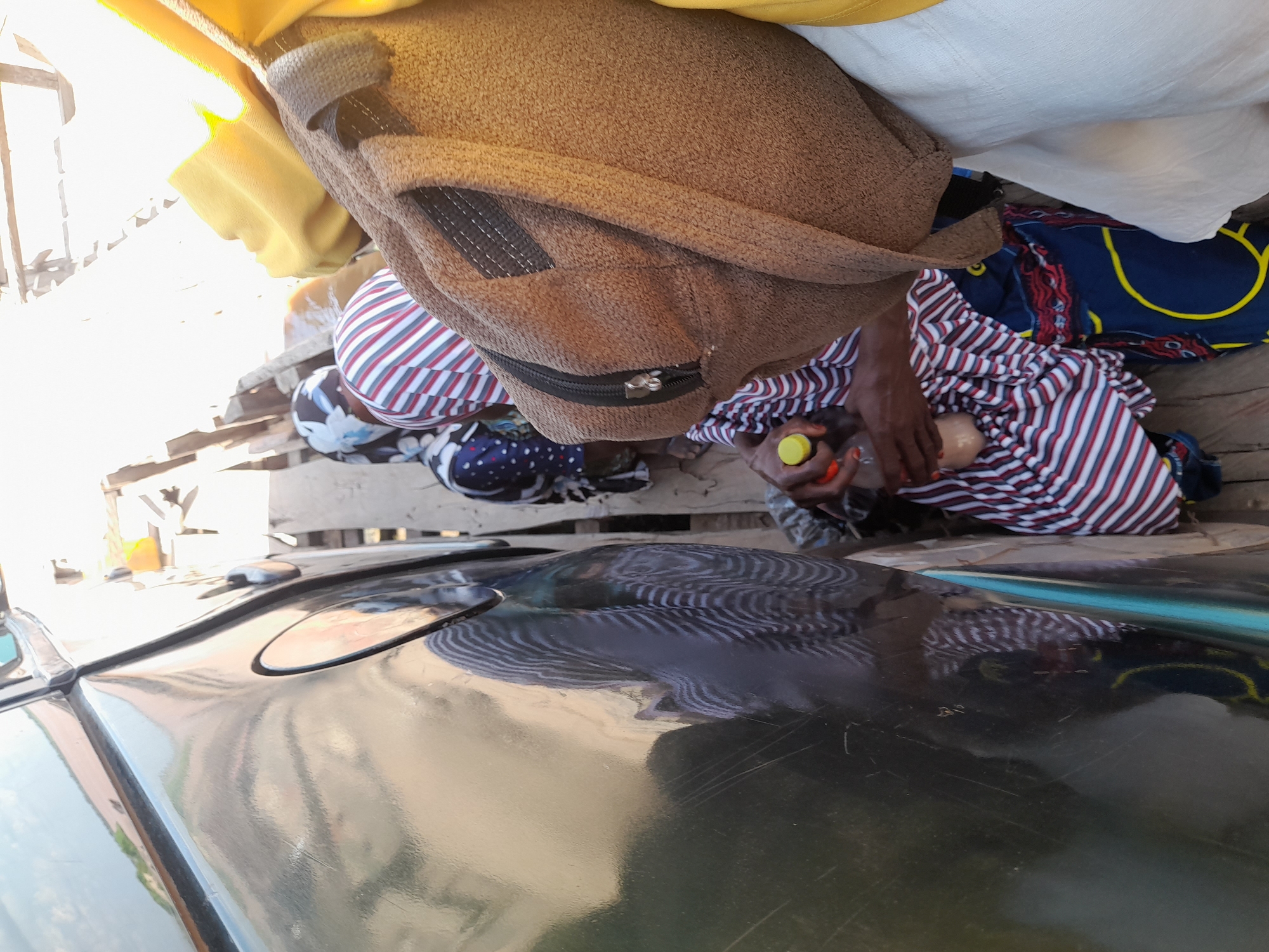 This image depicts a scene in the city of Ibi, Nigeria, likely on a wooden raft or boat. In the foreground, a person's hands are visible, holding a brown bottle with a yellow cap and another bottle containing a light-colored liquid. They are wearing a striped dress. To the right, a person is wearing a yellow top and a brown bag is prominently displayed. In the background, several other individuals are seen, some wearing headscarves. The environment appears to be outdoors, with natural light illuminating the scene, creating a bright atmosphere. The presence of a large, dark vehicle or object on the left side of the image suggests transportation or a substantial structure. The overall impression is one of a busy, possibly crowded, environment, consistent with daily life in a Nigerian city.