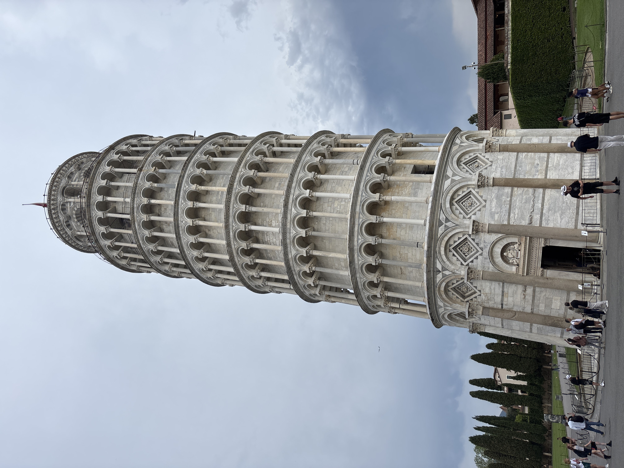 The image shows the Leaning Tower of Pisa, located in Pisa, Italy. The tower, built of white marble, is leaning to the right. There are people standing near the base of the tower, and tall, slender trees line the pathway leading to the tower. The sky is overcast with gray and white clouds. Part of a building covered with green vines is visible on the right side of the image.