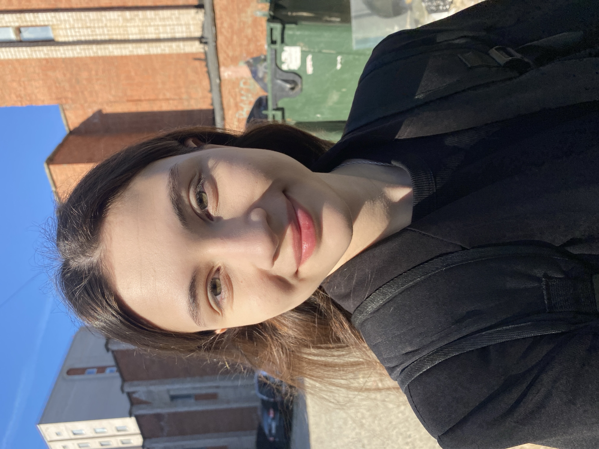 A young woman is posing for a selfie outdoors in Votkinsk, Russia. She is wearing a black jacket or backpack and has long brown hair. In the background, there are brick buildings and green garbage bins. The sky is blue.