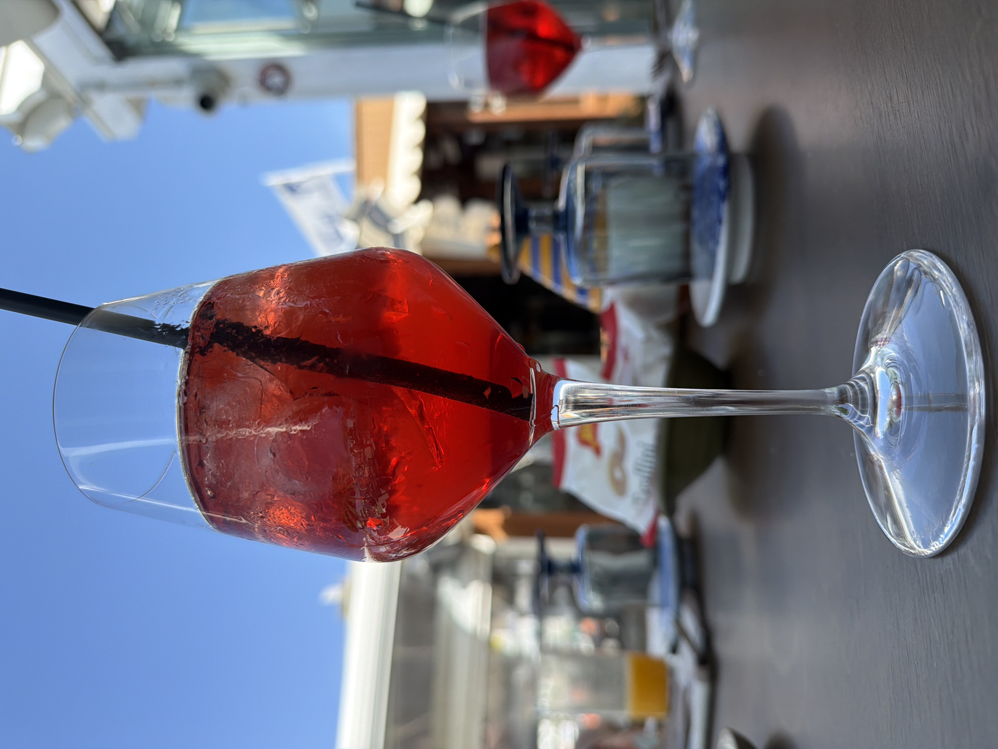 A close-up, medium shot depicts a glass of red liquid, possibly a cocktail, filled with ice and a black straw. The glass is sitting on a dark-colored, likely wooden, table. Behind the glass, another similar glass with a red liquid is visible, along with sugar packets and a glass with orange liquid. Background details suggest an outdoor setting. In the distance, architectural elements include awnings, a security camera, and glimpses of a clear blue sky. This photo was likely taken in Ischia, Italy.