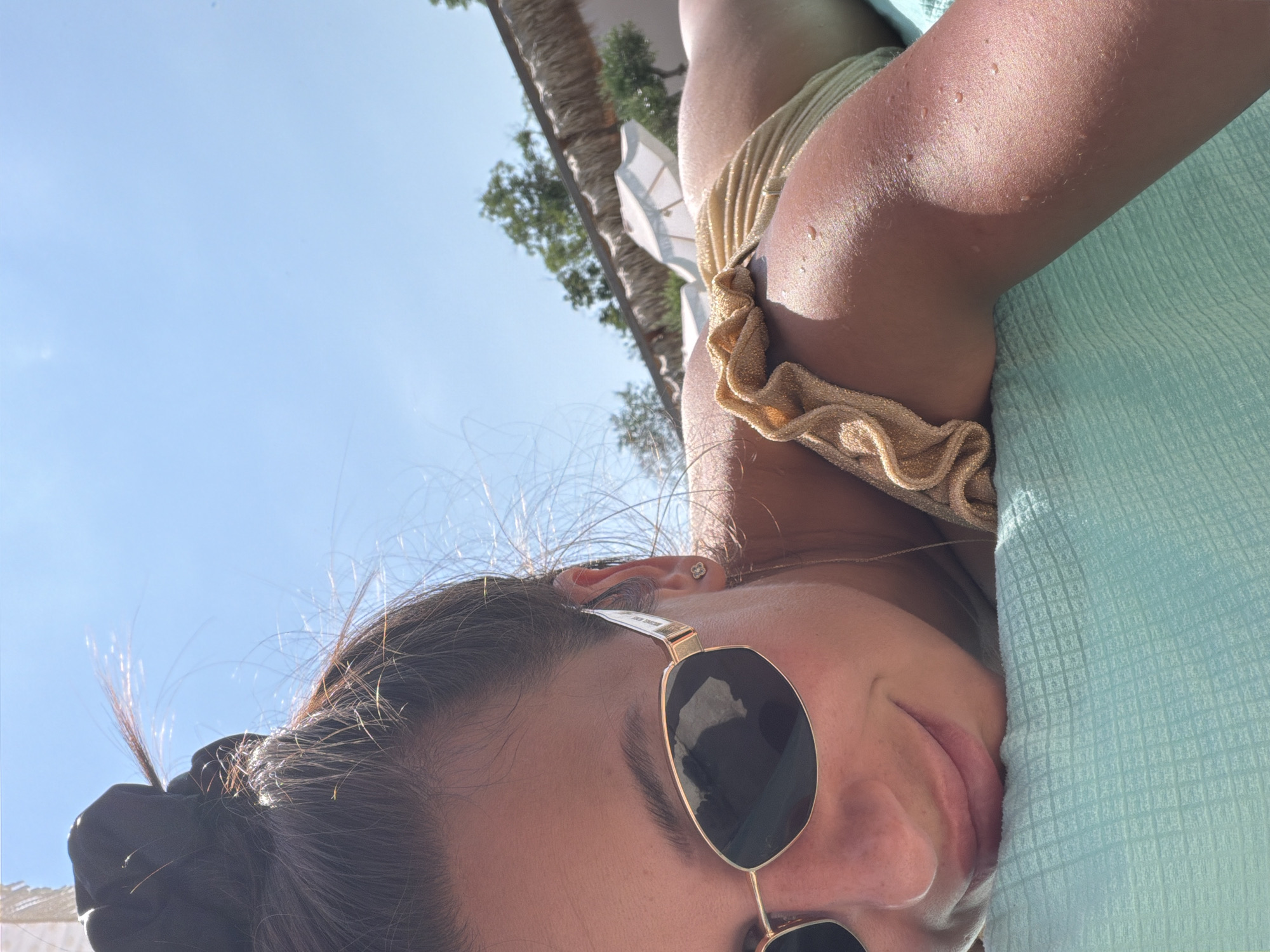 A young woman with brown hair pulled up in a bun is lying on a turquoise towel under a clear blue sky. She is wearing sunglasses and a gold bikini. Her skin is glistening with water droplets. In the background, there are trees, umbrellas, and a thatched roof structure. The scene suggests a sunny day at a resort or beach. The location is not specified.