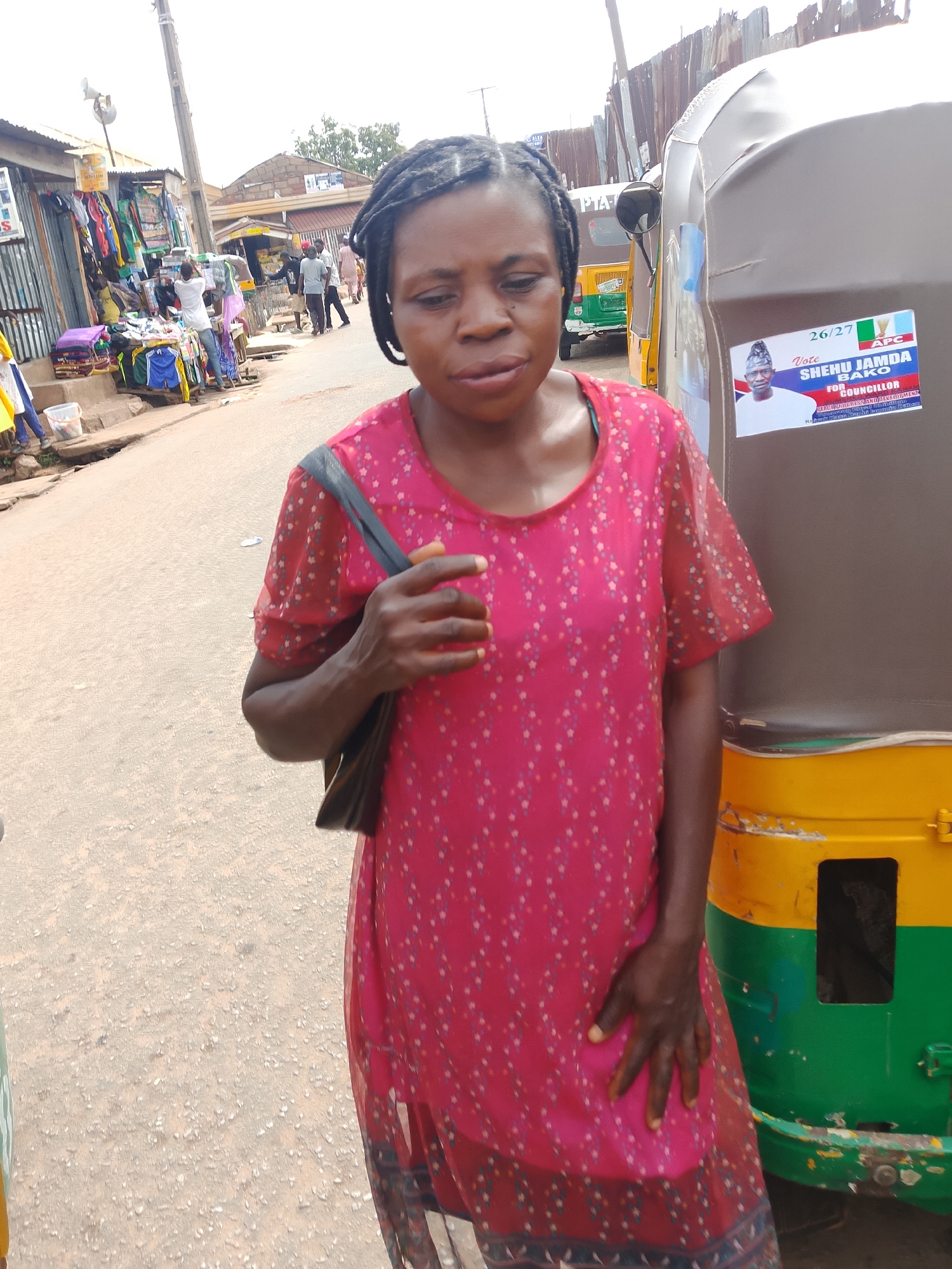 An outdoor, medium shot captures a Black woman standing next to a yellow and green auto-rickshaw in what appears to be a marketplace. The woman, facing slightly left, has dark skin and braided hair. She is wearing a short-sleeved, sheer, pink dress with a faint diamond pattern in shades of red, blue, and white. Her left hand rests on her stomach, and her right hand is visible at her side. A dark-colored bag is slung over her left shoulder.
The auto-rickshaw, a common form of transportation in many parts of the world, is positioned to the right of the woman. A political sticker with a picture of a man in a cap, along with text and the Nigerian flag, is adhered to its side. In the background, a row of stalls or shops lines a street, displaying various goods, including clothing. Several other people, some appearing to be shoppers or vendors, are visible in the distance, suggesting a busy commercial area. The ground is paved with asphalt or concrete, and a utility pole is visible on the left side of the frame. The sky is overcast, indicating a cloudy day.