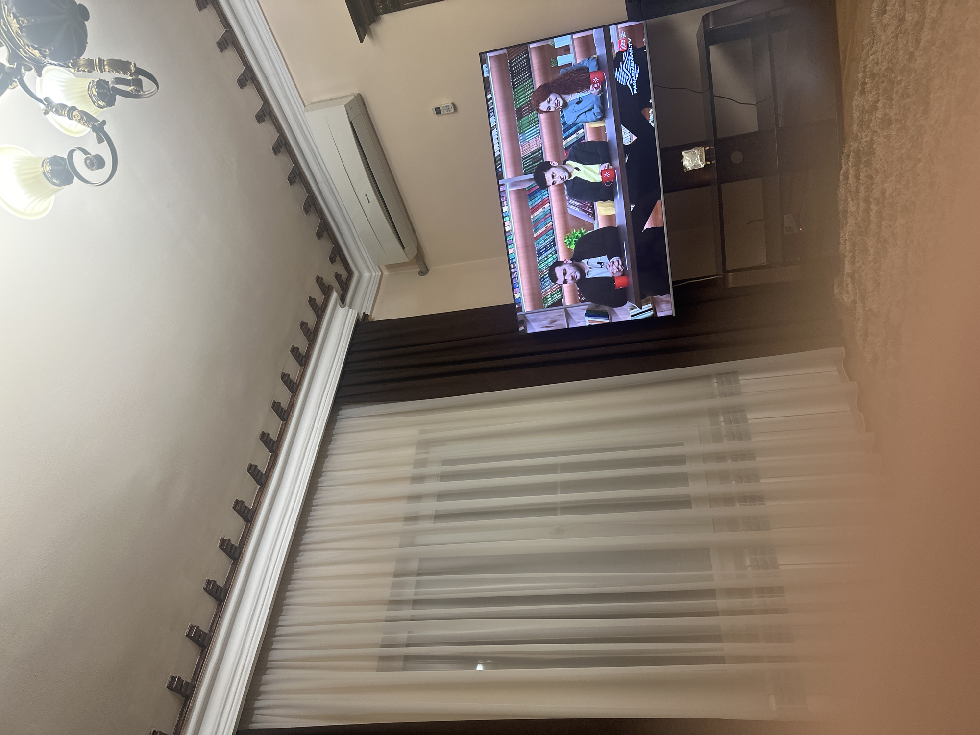 The image shows the interior of a room. A television screen is visible, displaying what appears to be a talk show, featuring three people seated at a table against a backdrop of shelves filled with books. The logo of "PANARMENIAN.TV" is displayed on the screen. The television is mounted on a wall above a window covered with sheer white curtains. The wall is painted in a light neutral tone and features decorative molding. In the upper left corner, a decorative chandelier hangs from the ceiling. Additionally, there is a light-colored air conditioning unit mounted on the wall near the television. Based on the appearance of the talk show shown on the television, the image is likely located in Yerevan, Armenia, as "Panarmenian TV" is an Armenian broadcasting network.