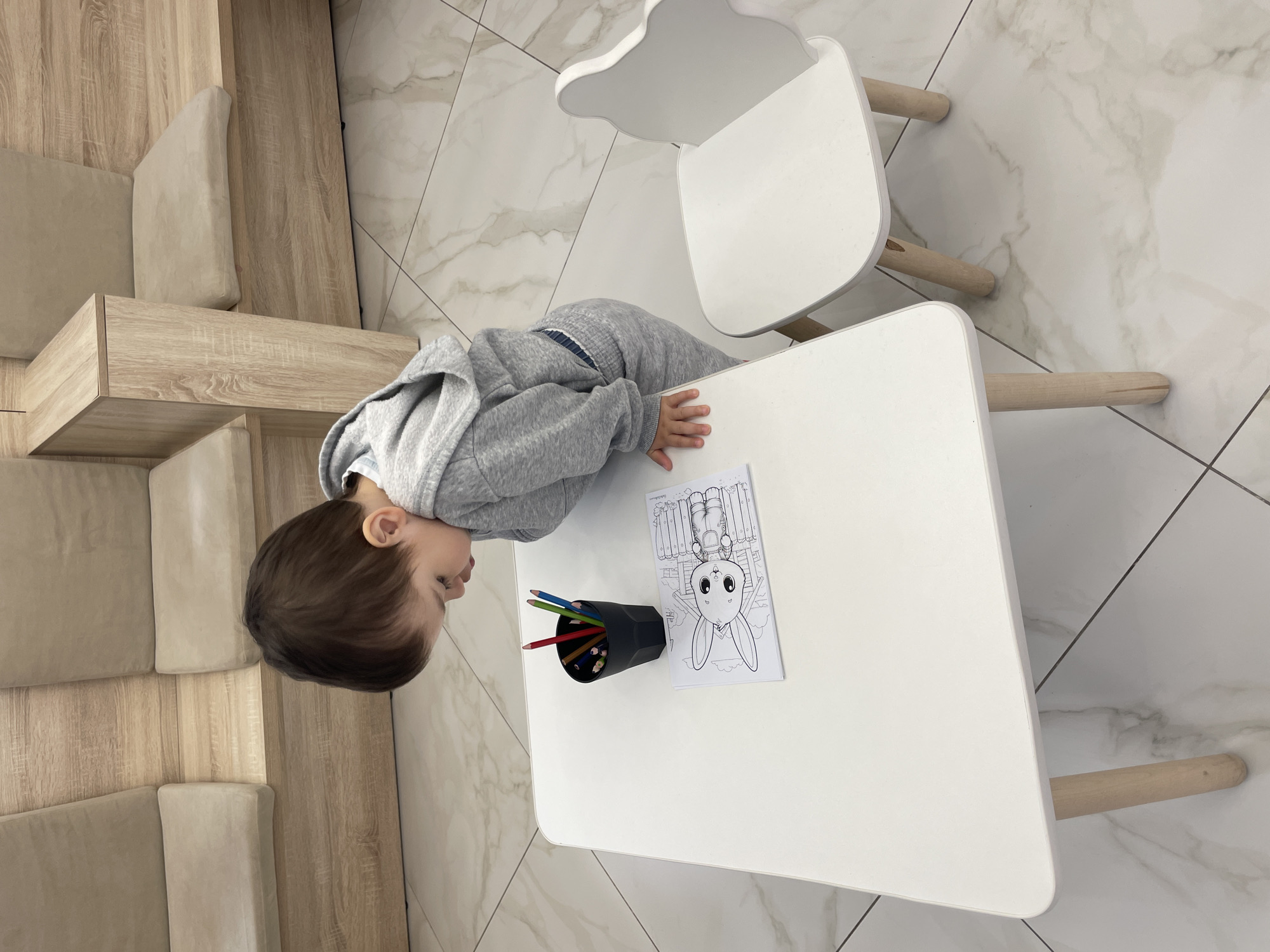 A young child is shown from an overhead perspective, leaning forward over a small white table. The child, likely a toddler, has short dark brown hair and is dressed in a grey hooded sweatshirt and matching grey trousers. Their left hand is resting on the table.
On the white table, a black hexagonal cup holds a collection of colored pencils. Next to the cup is a coloring page featuring a large outline of a bunny rabbit with a large head, along with other elements in the background that appear to be part of a fence or building. The child seems focused on the coloring page or the pencils.
To the right of the table, a small white chair with light wooden legs is visible. The floor throughout the scene is covered with light-colored tiles, possibly marble or a similar material, with subtle darker veining and dark grout lines. To the left of the child and table, there's a section of modern-looking furniture, consisting of light wooden panels and beige upholstered cushions, possibly part of a modular bench or seating area.
The scene depicts an indoor setting, likely a play area or a common room, with bright, even lighting. The child appears engaged in creative play. The location context is Izhevsk, Russia. No discernible text is visible other than the outlines on the coloring page.