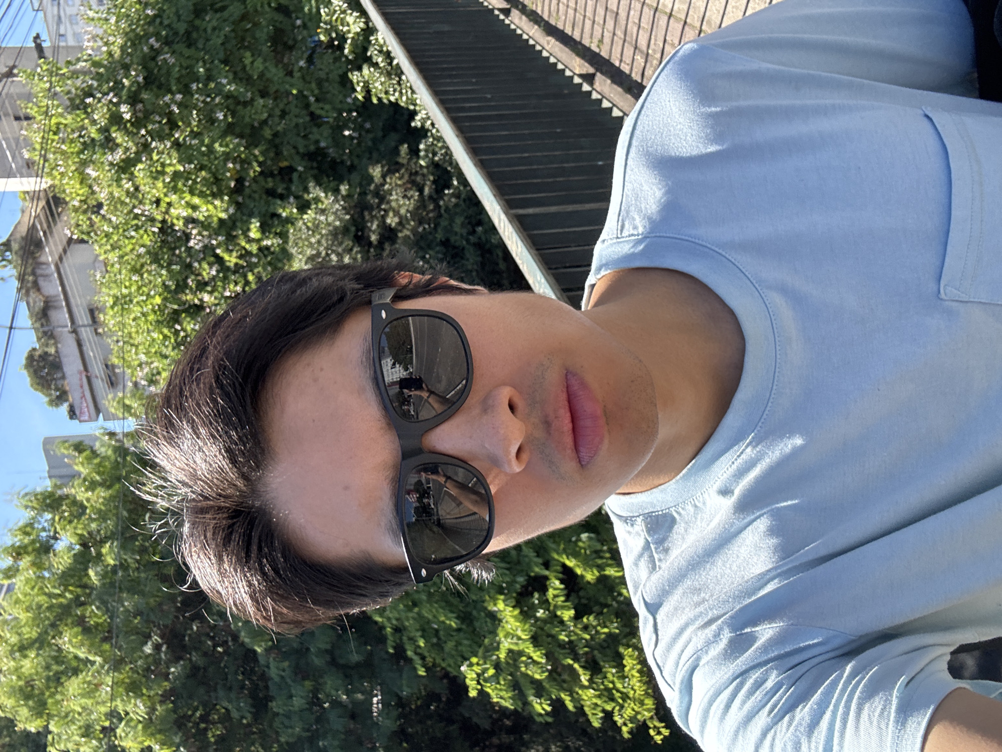 A man wearing sunglasses and a light blue t-shirt takes a selfie. He is likely on a pedestrian overpass, with trees and buildings in the background. The reflection in his sunglasses reveals a cityscape, possibly within São Paulo, Brazil. A pedestrian walkway with metal railings is to the right of him. The sky is clear and it is daytime.