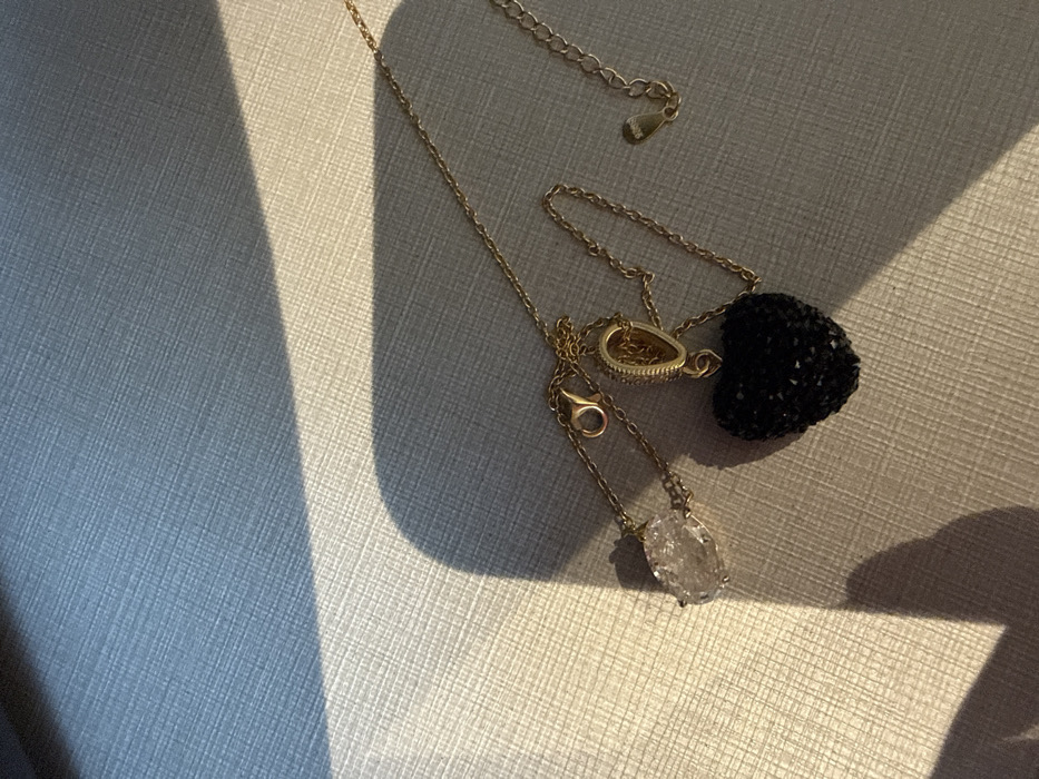 Two gold-colored necklaces are presented on a light-colored, textured surface exhibiting a crosshatch pattern, possibly fabric or Saffiano leather. Strong directional lighting casts a diagonal shadow across the upper left and lower left portions of the frame, illuminating the central and right areas.
The first necklace, positioned to the left, features a clear, irregularly shaped, faceted pendant, resembling a quartz crystal. Its chain includes a lobster claw clasp. The second necklace, located centrally, displays a black, heart-shaped pendant densely covered with small, reflective black crystals or beads. This heart pendant is attached to its gold-colored chain via a curved bail. This second chain also includes a lobster claw clasp and an extender chain with a small tag at its end. No individuals or active interactions are depicted. The specific location within Dubai, United Arab Emirates, is not discernible from the visual information provided.