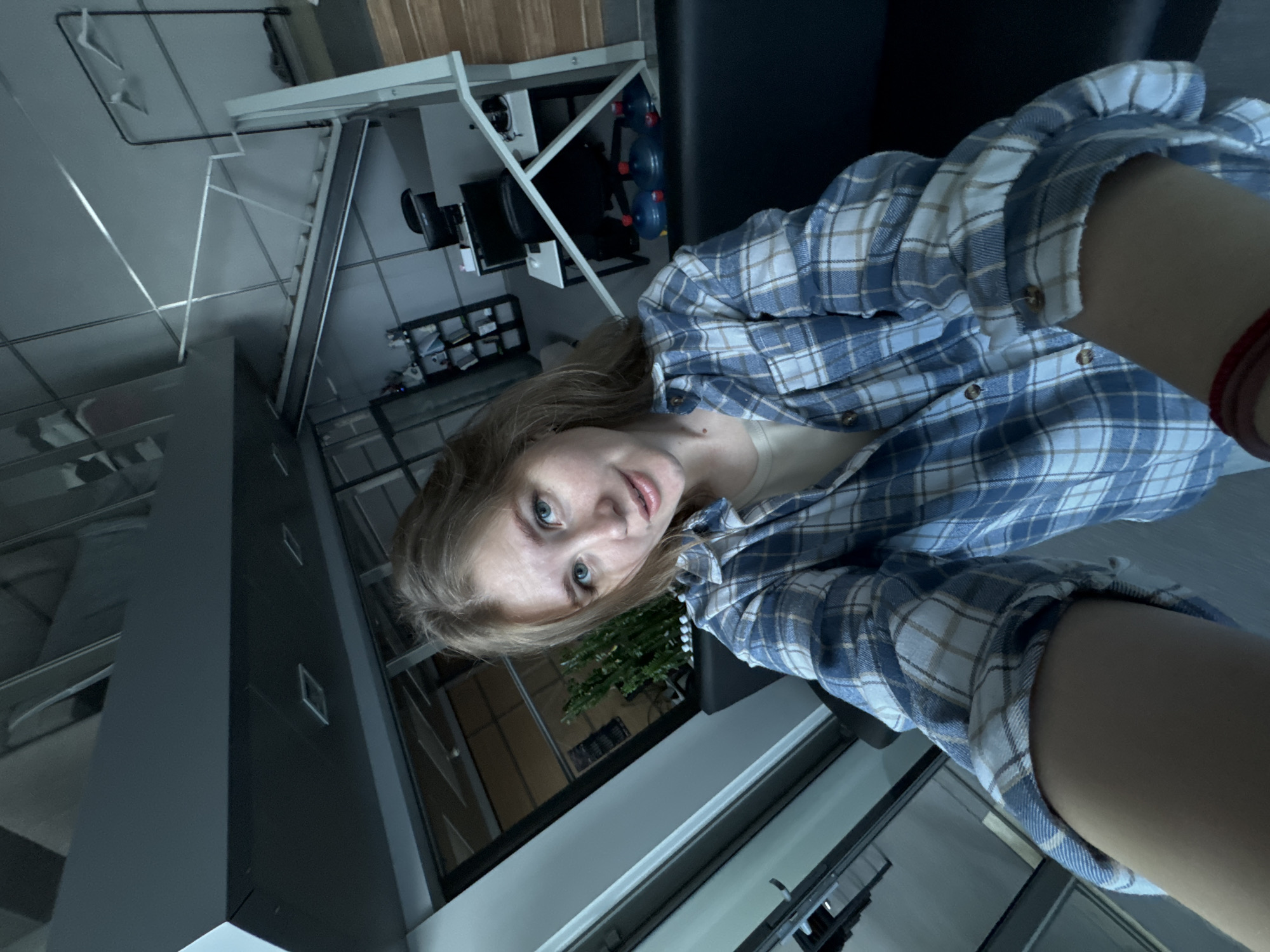 A young woman with blonde hair and blue eyes is taking a selfie in what appears to be an office space. She is wearing a blue and white plaid shirt over a beige top. The background shows modern office furniture, cubicles, shelves with supplies, and a large window with a view of an outdoor area that includes a plant. The image is taken from a high angle, looking down at the woman. The location of Perm, Russia, cannot be determined from the visual information provided in the image.
