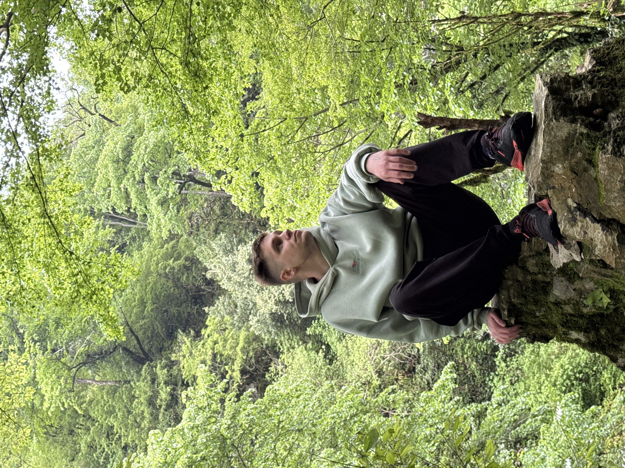 A young man is sitting on a moss-covered rock outcropping in Tsabal, Abkhazia, with a dense forest of green foliage as a backdrop. He is wearing a light green hoodie, dark pants, and dark sneakers with red accents. He is looking upwards, seemingly towards the sky or treetops.