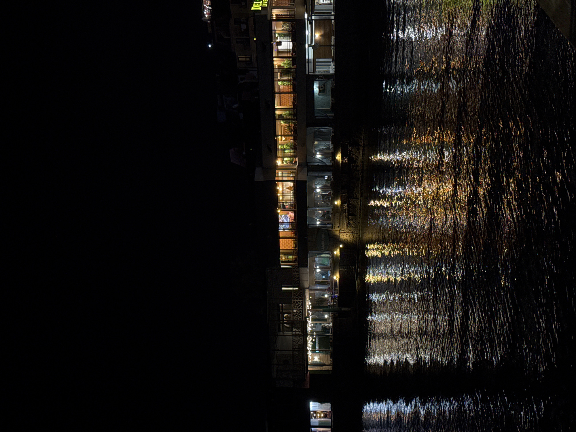 The image shows a building illuminated at night, with its lights reflecting on the water in front of it. The building appears to be a restaurant or a complex of establishments, with visible windows, signs, and decorative lighting. The architecture suggests a modern design, possibly with multiple levels. The water is dark and choppy, with the reflections creating streaks of yellow and white light. The sky is completely black, indicating nighttime. In the upper right portion of the image, a sign with the word "DEL" is visible, followed by smaller text that is partially obscured. Based on the prompt's context, this scene is located in Pomorie, Bulgaria. There are no visible people or specific actions taking place.