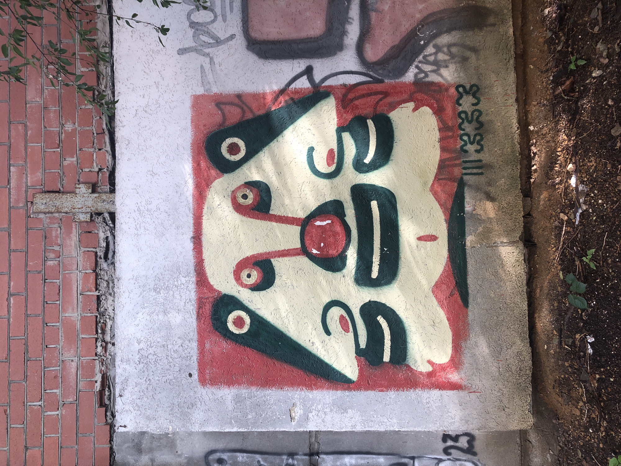 The image shows a graffiti artwork on a concrete wall in Yekaterinburg, Russia. The graffiti depicts a stylized, cartoonish face with large eyes, a red nose, and green accents. The face is predominantly yellow with red and green outlines and details. The concrete wall is textured and appears to be part of an outdoor structure, possibly a building exterior or a retaining wall. Above the concrete wall is a brick wall made of red bricks. The ground in front of the wall is covered with dirt, small plants, and debris, suggesting an urban outdoor environment. There are other faint graffiti tags on the right side of the wall. The overall scene appears to be a public space in Yekaterinburg where street art is present.