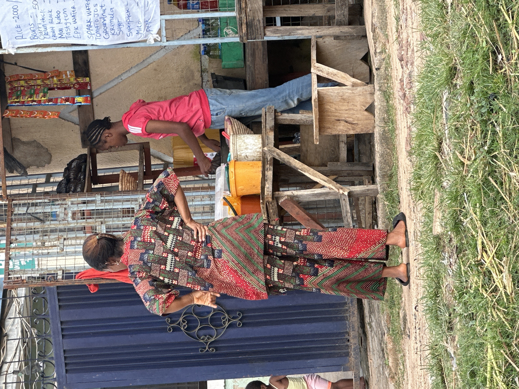 In Narayi, Nigeria, a woman in a vibrantly patterned dashiki and pants stands with her hands on her hips, facing away from the camera towards a stall. She is positioned in front of a dark blue metal gate. To her right, a young girl wearing a pink hooded sweatshirt and blue jeans is attending to the stall. The stall itself is made of wood and metal and features shelves displaying various items, including bottles of drinks and packaged goods. A handwritten price list is affixed to a board. In the foreground, dry grass and dirt are visible. A smaller child in a light-colored top and pink shorts can be seen to the far left, near the gate.