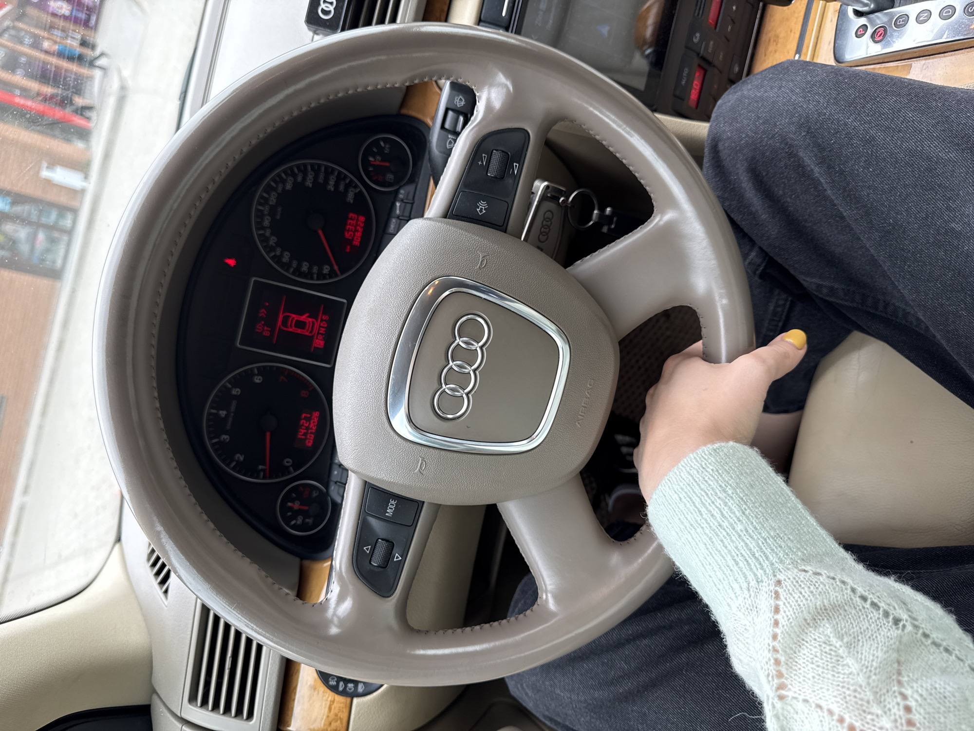 The image shows a close-up of the interior of an Audi A4 (B7) sedan, viewed from the driver's perspective. The steering wheel, dashboard, and part of the center console are visible. A person's hand, wearing a light blue knitted sweater, is resting on the steering wheel. The dashboard displays the time as 14:27 and the odometer reading as 15333. The car is likely stationary, as the gear selector is visible in the "P" (Park) position. The exterior environment, seen through the windshield, suggests an urban setting with a brick building and shops. The lighting indicates it is daytime. The specific location within Yartsevo, Russia, cannot be determined from the visual information provided.