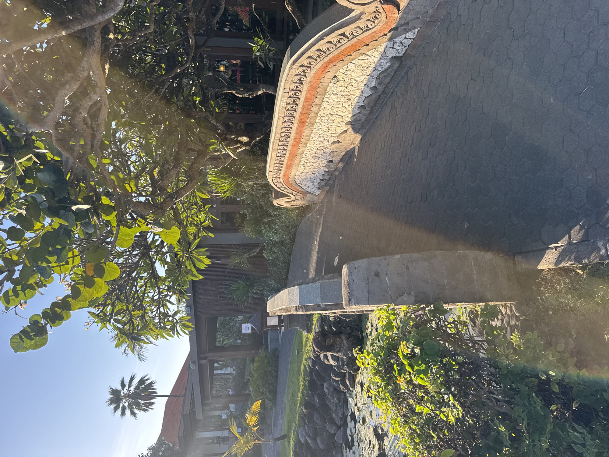 An outdoor shot captures a picturesque scene in Kuta Selatan, Indonesia, featuring a curved bridge with ornate railings leading towards a building complex. The bridge's railings are adorned with a mosaic of white stones and a decorative band of red and carved patterns. The walkway of the bridge is paved with hexagonal stones. To the left of the bridge, a serene pond is visible, populated with water lilies and bordered by a rocky embankment covered in lush greenery. Beyond the pond, a low-lying building with a red-tiled roof and a modern facade is situated. Tropical trees, including a tall palm tree, are scattered across the landscape, their leaves catching the warm sunlight. The overall atmosphere is tranquil and inviting, with the bright sky and dappled sunlight enhancing the natural beauty of the location.