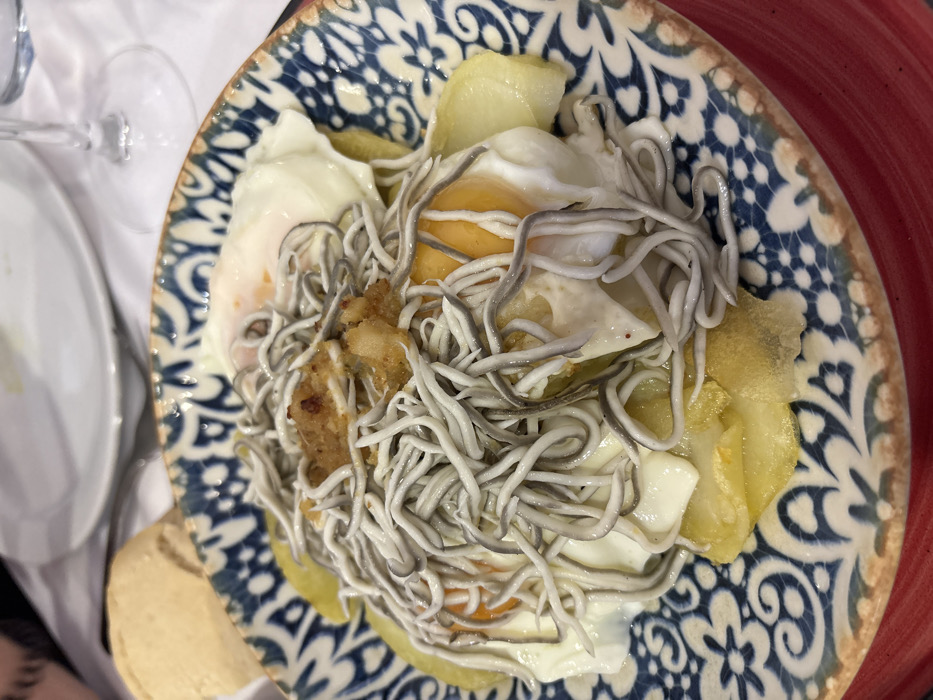 The image displays a close-up of a prepared dish served in a restaurant in Córdoba, Spain. The central subject is a round ceramic plate with a distinctive blue and white patterned rim, resting on a larger red-rimmed plate. The dish consists of several fried eggs with intact, bright orange yolks partially obscured by white albumen, placed over a bed of thinly sliced, pan-fried potatoes. Abundantly layered on top of the eggs and potatoes are numerous slender, greyish-white strands resembling *gulas* (imitation baby eels). A small amount of browned, textured pieces, likely fried garlic or pepper, are distributed among the *gulas*. In the background, out of focus, a light-colored bread roll, a white tablecloth, a clear stemmed glass (likely a wine glass), and a portion of another white plate are visible, indicating a dining establishment. The precise location within the city of Córdoba cannot be determined from the visual information provided.