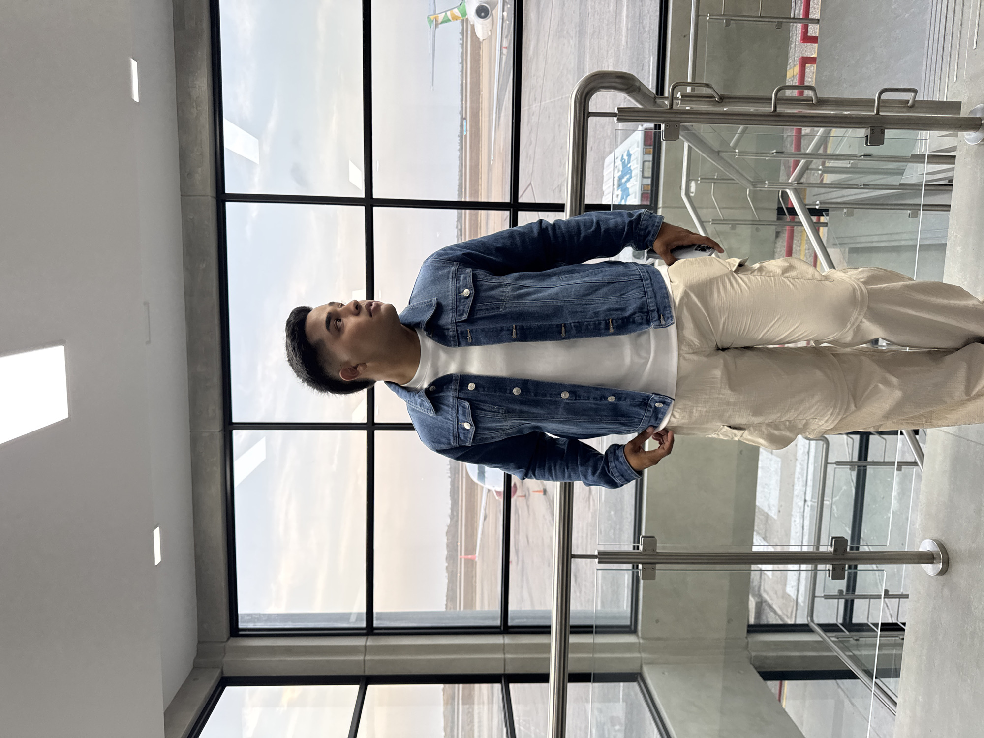 A man is standing inside an airport terminal in Cotoperíz, Venezuela. He is wearing a white t-shirt, a denim jacket, and light-colored pants. He is looking up and to his left. Behind him is a large window with a view of an airplane on the tarmac. To his right are metal and glass railings. The environment appears modern, clean and well-lit. He is holding a black object in his right hand.