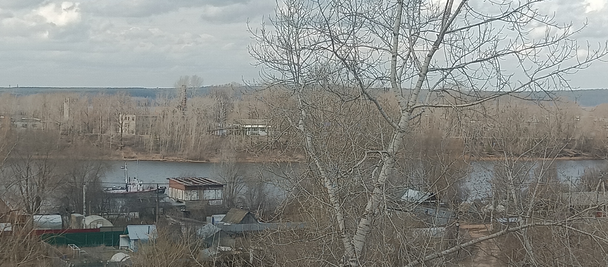 This image captures a wide, somewhat hazy view of a landscape featuring a body of water, distant land, and foreground elements.
In the foreground, the branches of several bare, leafless trees dominate the right and upper portions of the frame, indicating late winter or early spring. Their intricate network of branches is a prominent visual feature. Below these trees, on the near bank, glimpses of small, perhaps residential or storage structures with fences are visible.
A calm, dark blue-grey body of water, likely a river, stretches across the middle of the image, separating the foreground from the distant bank. On the far bank, a dense line of bare trees covers the terrain. Scattered amongst these trees are several buildings. To the left, an old, tall industrial-like structure with a chimney stands out. Further right, another multi-story building, possibly residential or administrative, is visible. Near the left side of the far bank, a boat, appearing to be a small tugboat or work vessel, is moored on the water. Another smaller structure with a reddish roof is near the boat on the water's edge.
The horizon in the distant background shows a faint, undulating treeline under a heavily overcast sky, with uniform grey clouds suggesting a muted, diffused light. No direct sunlight is visible. The overall impression is one of quietude and perhaps a slightly melancholic atmosphere due to the bare trees and cloudy weather. There is no discernible human activity or legible text.