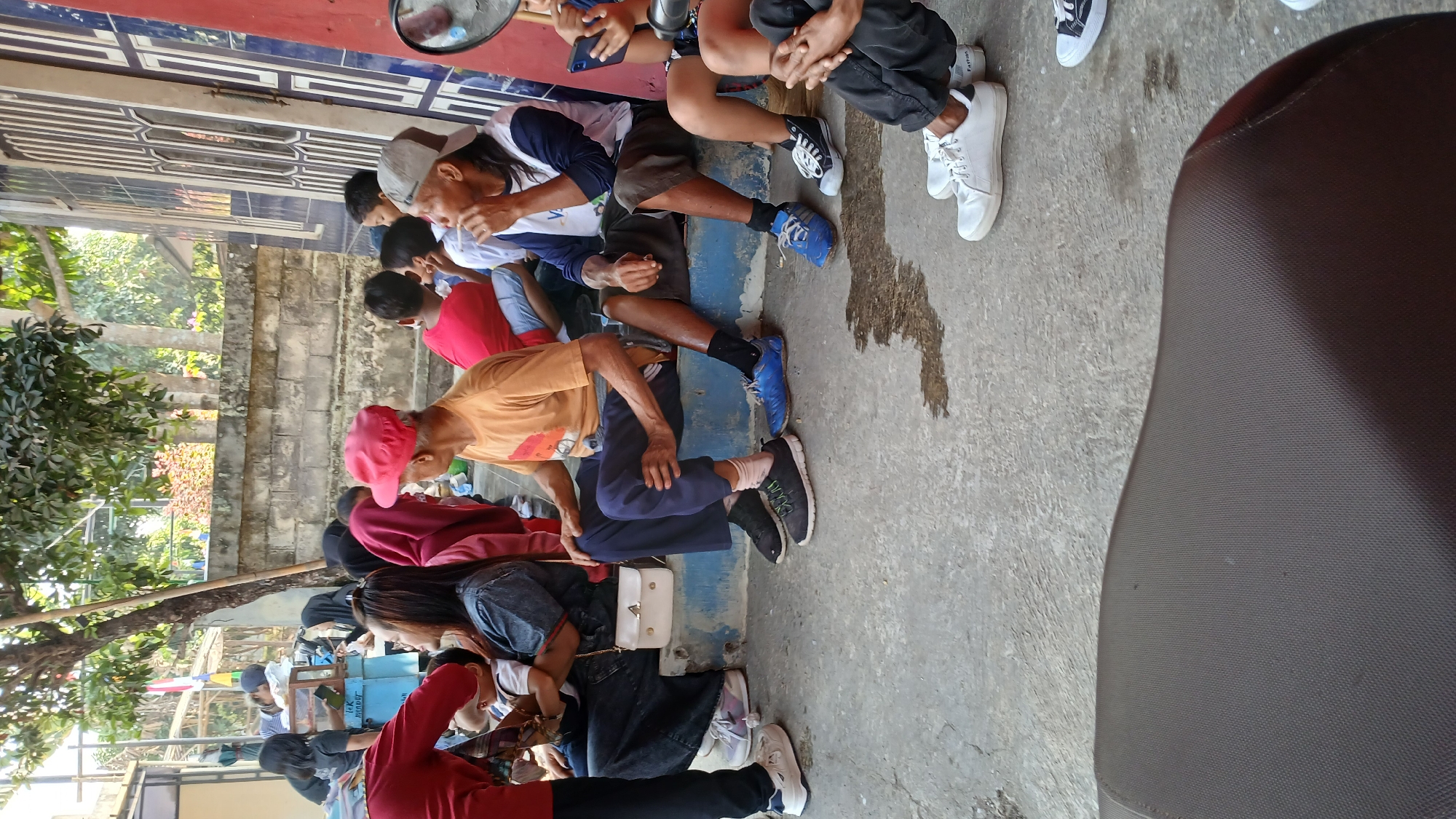 In Mendut, Indonesia, a group of people are gathered outdoors on a concrete ground in front of a building with blue and white architectural elements. The scene appears to be a communal gathering or waiting area.
In the foreground, the seat of a motorcycle or similar vehicle is visible, taking up the bottom portion of the frame.
Further back, several individuals are seated on a raised blue ledge. On the far left, a person in a red shirt and black pants is partially visible, facing away from the camera. Next to them, a woman in a dark denim top is holding a baby. To her right, a person wearing a maroon hoodie is seated.
The central figure of the image is an elderly man wearing an orange t-shirt and a red cap, seated with his legs crossed. He appears to be looking to his left. Beside him, a man wearing a grey baseball cap, a blue and white shirt, and brown shorts is seated, holding something to his mouth, possibly a cigarette or a pen.
Behind these seated individuals, several children and adults are visible, some in red shirts. The background features a brick wall and lush green trees, suggesting an outdoor setting with a residential or community atmosphere. The overall impression is one of casual interaction and shared space in a neighborhood.