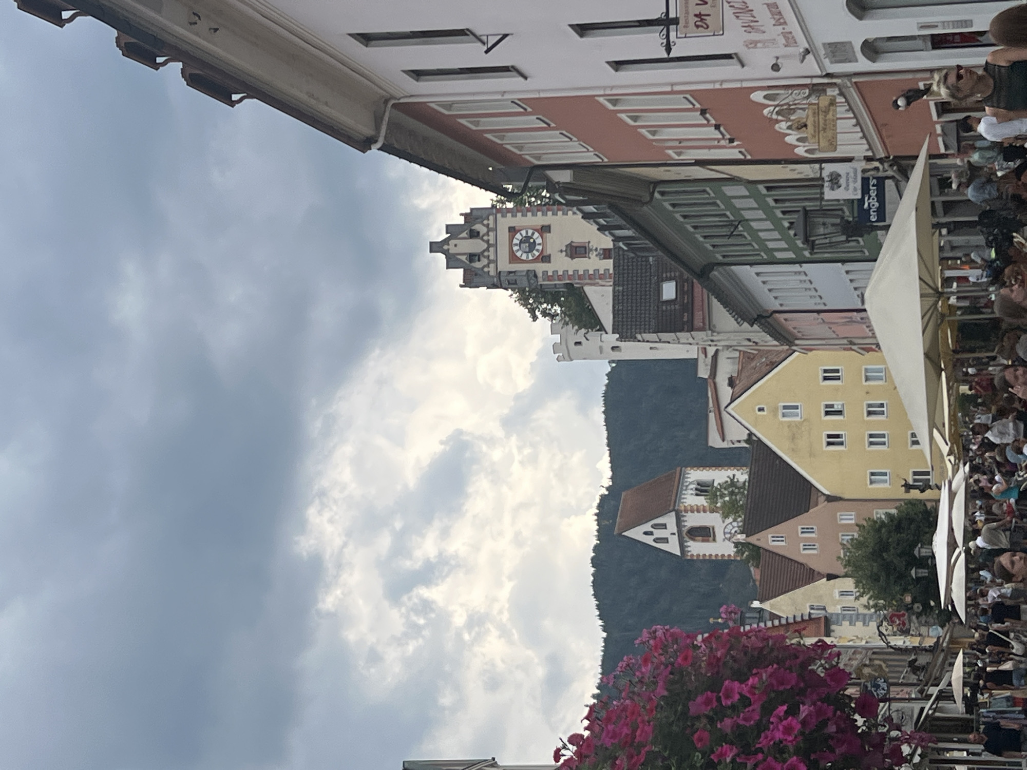 The image depicts a street scene in Füssen, Germany, characterized by historic architecture and a bustling atmosphere. In the foreground, a vibrant display of pink petunias cascades from hanging baskets, adding a splash of color to the scene. The street is lined with multi-story buildings, displaying a variety of architectural styles and muted color palettes, including shades of white, pink, yellow, and green. Prominent architectural features include gabled roofs, numerous windows, and decorative trim.
Dominating the background are two distinct towers: a clock tower with a large circular clock face and a white, crenellated tower. These structures are characteristic of medieval European architecture, likely part of the city's historic fortifications or religious buildings. Behind the towers, a dark, tree-covered mountain range serves as a backdrop, suggesting the city's scenic location.
The street is populated with a large number of people, indicating a lively commercial or tourist area. Individuals are seen walking, some carrying bags, and others gathered around shops or cafes. Outdoor seating with umbrellas suggests the presence of restaurants or cafes catering to the patrons. Several signs are visible on the buildings, some advertising businesses like "engbers" and "SANTINCI PIZZA - RESTAURANT." The sky is overcast with heavy, grey clouds, suggesting a potentially damp or cool day. The overall impression is one of a well-preserved historic European town center with active commerce and pedestrian traffic.