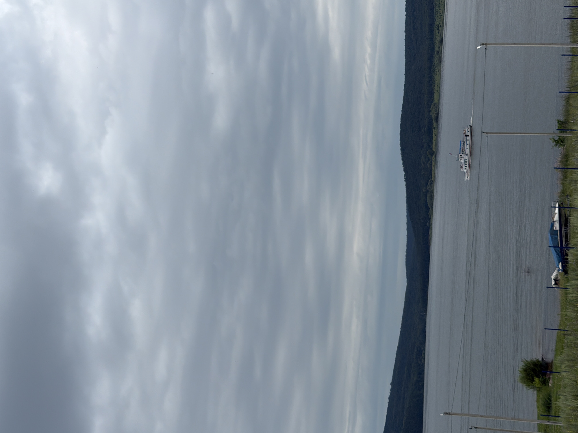 A wide, gray river dominates the lower half of the frame, reflecting an overcast sky filled with textured clouds. On the far bank of the river, a dark, densely forested hillside slopes upwards, its peak shrouded in the hazy atmosphere. In the middle distance, a white passenger boat with blue accents travels from left to right, leaving a wake on the water. The foreground features a grassy embankment with several utility poles and wires running horizontally across the scene. On the embankment, a blue tarp covers at least one boat. The location is the city of Sergeevka, Russia.