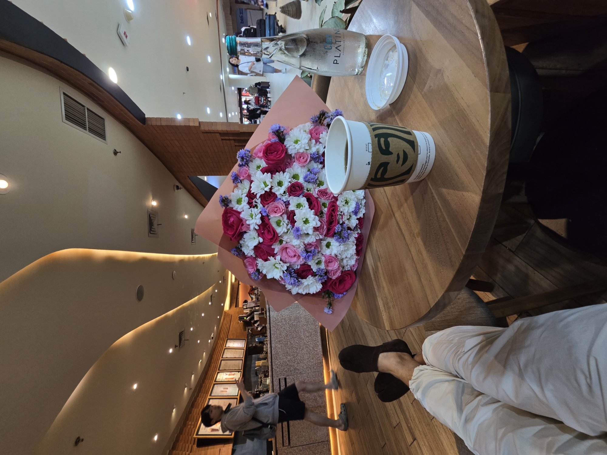 The image captures an indoor scene from a first-person perspective, focused on a wooden round table where a vibrant bouquet of flowers, a Starbucks cup, and a bottle of sparkling water are placed. The bouquet, prominently featured, consists of pink and white roses, white daisies or chrysanthemums, and small purple flowers, all wrapped in a pink paper cone. Next to it, a Starbucks paper cup with its green logo shows partial text including "DECAF" and phrases like "ge you're about to enjoy" and "transfer. Avoid contac". A clear glass bottle of "Clea PLATINUM Sparkling Water" with a green cap and a white plastic lid rests beside the cup.
The person taking the photo is seated, with their legs visible in light-colored trousers and dark sandals/slippers, crossed at the ankles.
The setting appears to be a modern cafe or a section within a mall in Kuta, Indonesia. The background features curved, light-colored walls with warm accent lighting and recessed spotlights, interspersed with wooden architectural details and air vents. In the mid-ground, a person wearing a grey hoodie and dark shorts is seen walking, with other blurry figures indicating a public and active space. Further in the distance, what appears to be a service counter or display area is visible, along with potential luggage or shopping bags, suggesting a commercial or travel-related environment typical of tourist areas. The overall ambiance is relaxed and contemporary.