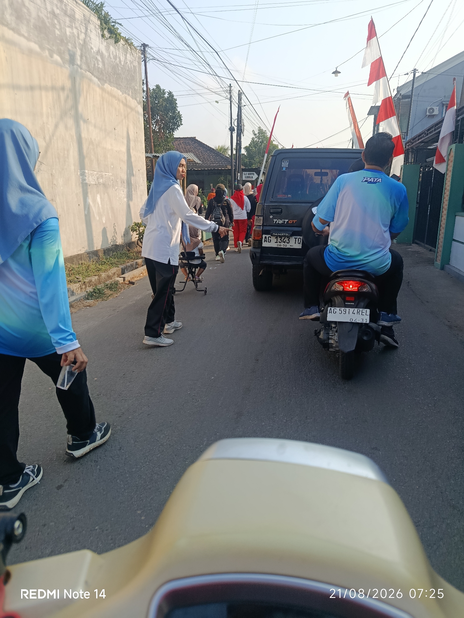 The image depicts a street scene in Balerejo, Indonesia, on August 21, 2026, at 07:25. Several individuals are present, some wearing headscarves and athletic attire, suggesting participation in an outdoor activity or event. Two women in blue headscarves are in the foreground, with one holding a plastic cup. Behind them, other people are walking along the paved road.
Dominating the mid-ground are two vehicles: a dark-colored SUV with the license plate "AG 1343 TQ" and a motorcycle with a rider wearing a blue and white shirt, displaying the license plate "AG 5914 REL". The motorcycle's brake light is illuminated. Indonesian flags, red and white, are visible on poles along the street, indicating a national celebration or observance. The environment appears to be a residential or commercial street with buildings on the right and a wall on the left. Overhead, power lines crisscross the sky. The overall atmosphere suggests a community gathering or a morning procession.