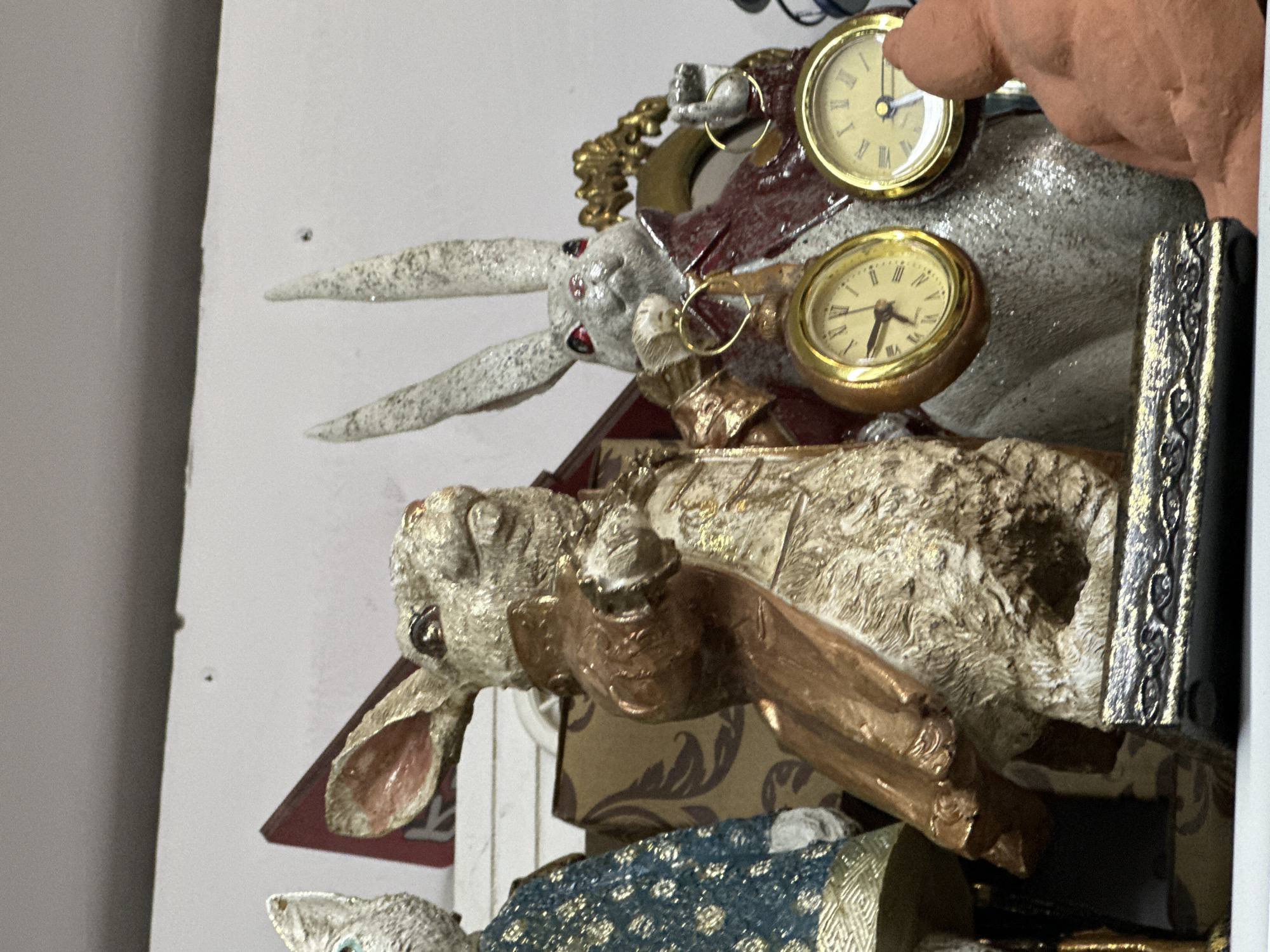 The image shows two decorative rabbit figurines, styled after the White Rabbit from Alice in Wonderland. The rabbit on the left wears a bronze-colored coat and is positioned in a dynamic pose, with its right arm extended. The rabbit on the right wears a maroon coat and holds two pocket watches, one in each hand. The figurines are placed on a shelf, with other decorative items visible in the background. There is no discernible location in the city of Sochi, Russia, or any indications of an outdoor environment. The scene is a still life arrangement of these collectible figurines.