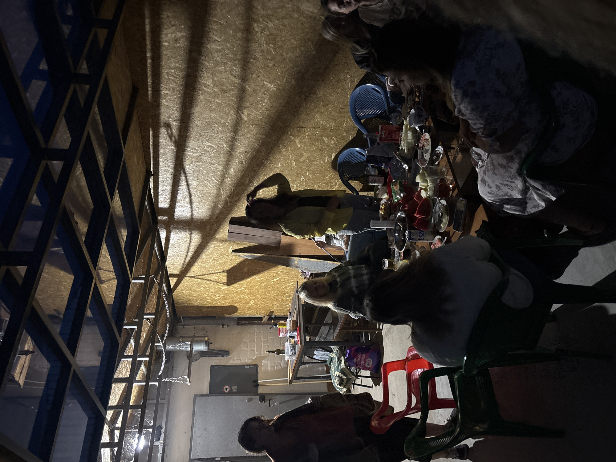The image depicts an indoor gathering of approximately eight individuals, likely at a casual event. The setting appears to be a semi-finished or industrial space, with exposed metal framing for a ceiling, unfinished wooden walls, and a concrete floor. One wall is a large, corrugated metal door.
Several people are seated around a large table laden with food and drinks, including watermelon slices, various dishes, bottles, and cups. Others are standing or moving around the periphery. One woman in a yellow jacket is standing near the wall, with her hand on her head. Another woman in a dark top is partially visible on the left, and a woman in a patterned dress is seated on the right, facing away from the camera. Two red plastic chairs and two blue plastic chairs are visible, along with green plastic chairs.
The lighting is dim and appears to be artificial, casting long shadows on the walls. There is a metallic chain hanging from the ceiling structure, possibly related to an industrial fixture or decoration. The overall atmosphere suggests a social gathering, possibly a barbecue or informal party. The location is identified as Chaikovsky, Russia, though no specific landmarks or street views confirm this within the image itself.