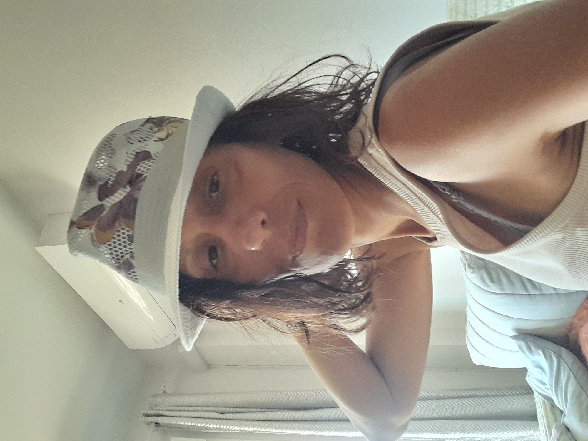 A female subject, with dark, shoulder-length hair, is depicted indoors within a room, identified as being in Manilva, Spain. She is wearing a light-colored, ribbed tank top and a light-colored fedora-style hat with a mesh crown featuring a brown and white pattern. A small ring is present in her right nostril. Her left arm is raised and bent, with her hand positioned behind her head, revealing a small, dark linear tattoo on her left forearm. Her right shoulder and upper arm are prominent in the foreground. The background consists of a light-colored wall, an upper-mounted white split-unit air conditioning system, and light-colored patterned curtains on the left. A light-colored, vertically ribbed piece of furniture, possibly a sofa or bed frame, and a light blue cushion are partially visible in the lower right background.