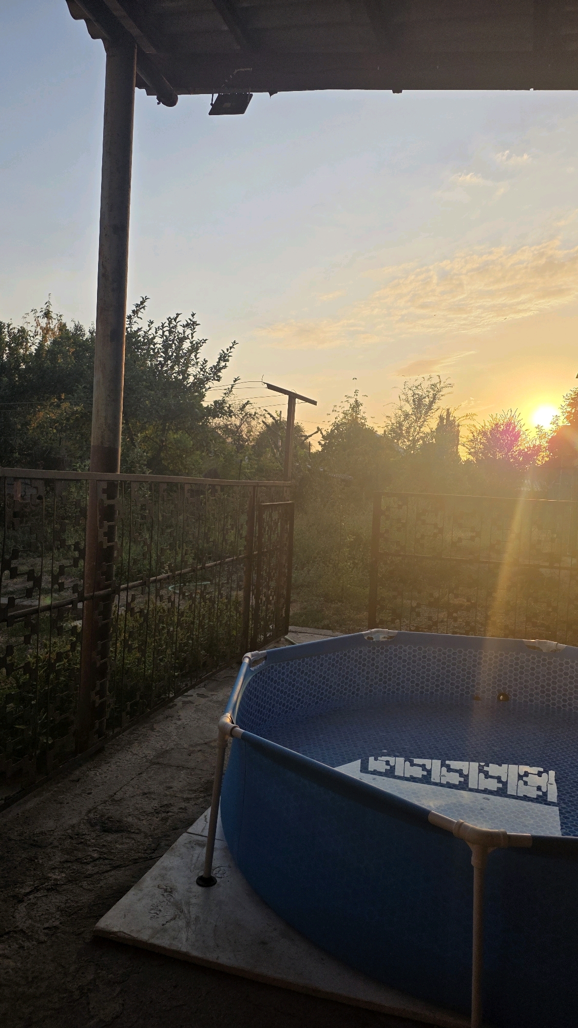 The image depicts a backyard scene during sunset, featuring an empty inflatable pool. The pool is blue with a hexagonal pattern on the interior and is supported by a light-colored frame. It rests on a concrete slab. To the left of the pool is a concrete patio and a metal fence. Beyond the fence, there are trees and vegetation. A large, weathered wooden post stands near the fence, supporting a roof overhang. A light fixture is attached to the underside of the overhang. The sky is partly cloudy with a gradient of orange and yellow hues, indicating the setting sun. The sun's rays are visible, creating lens flare effects across the image. The overall environment suggests a residential outdoor space, likely in a rural or suburban area given the surrounding vegetation and fencing. The location is specified as the city of Chuy, Kyrgyzstan.